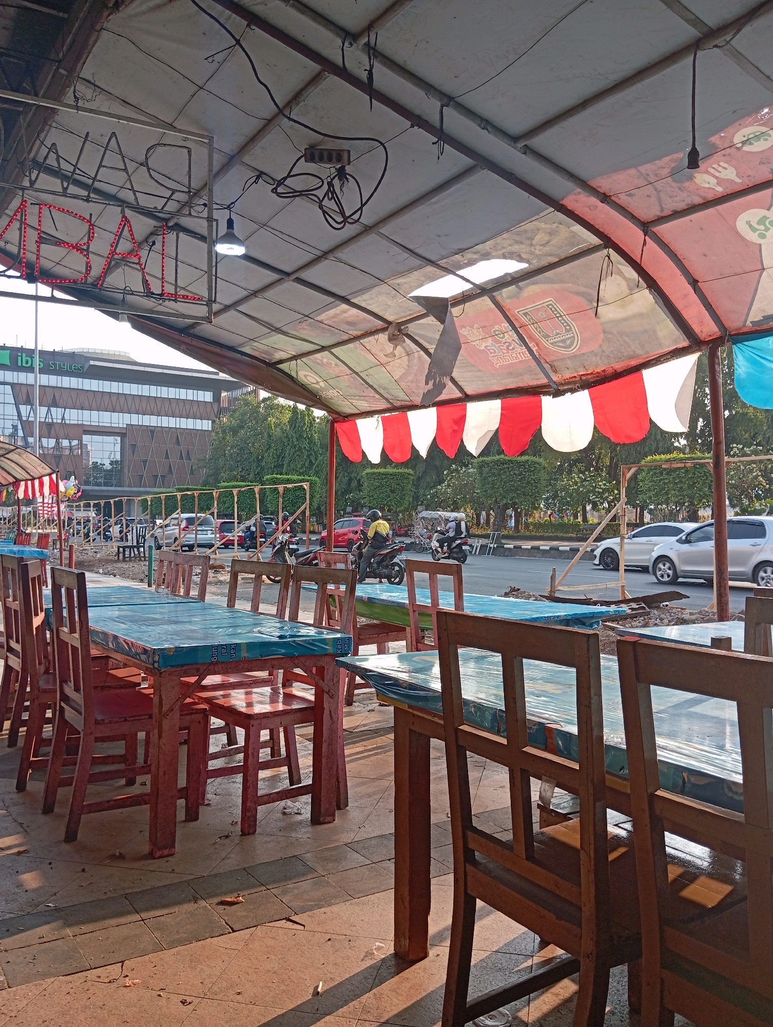 The image depicts an outdoor seating area of a roadside eatery in Semarang, Indonesia. The foreground is dominated by several wooden tables covered with blue plastic sheeting, with matching wooden chairs arranged around them. Some tables are empty, while others appear to have people seated, though their faces are not clearly visible. The tables are set on a tiled pavement, with shadows cast by the tables and chairs indicating the presence of sunlight.
Behind the seating area, a canopy made of translucent plastic and fabric with red and white stripes provides shade. Suspended from the canopy's metal frame are electrical wires and a bare light bulb. A neon sign with red lettering that appears to say "MIE AYAM" (Chicken Noodles) is visible, illuminated by red LED lights.
In the middle ground, a street is visible with several vehicles, including cars and motorcycles, in motion. A person in a yellow jacket is seen riding a motorcycle. On the sidewalk across the street, there is a row of trees and a modern building with a distinctive patterned facade, identified by a sign as "ibis STYLES." The overall scene suggests a busy urban environment where a local eatery operates alongside city infrastructure. The presence of a building labeled "ibis STYLES" strongly indicates the location is near an Ibis Styles hotel. Considering the typical urban layout in Indonesia, this scene likely portrays a street-side food stall operating in a commercial or mixed-use area within Semarang.