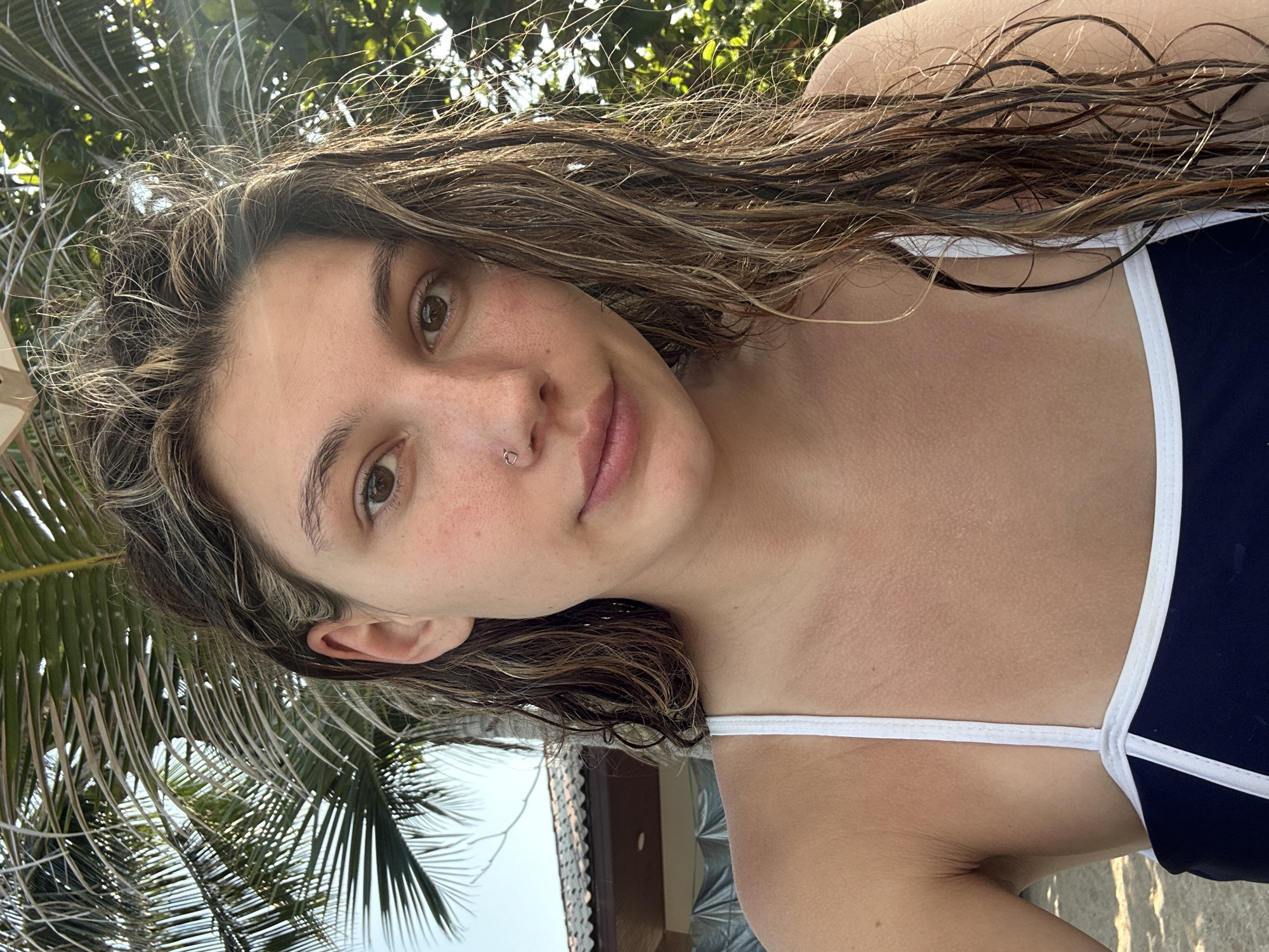 A woman with wet, brown hair is the primary subject of this outdoor photograph taken in Baan Chaweng Noi, Thailand. The woman is wearing a dark-colored swimsuit with white straps. She has a piercing in her left nostril and is looking directly at the camera with a slight smile. The background consists of palm trees and a building with a tiled roof, suggesting a tropical or coastal environment. The photograph appears to be taken from a low angle, looking up towards the subject and the sky.