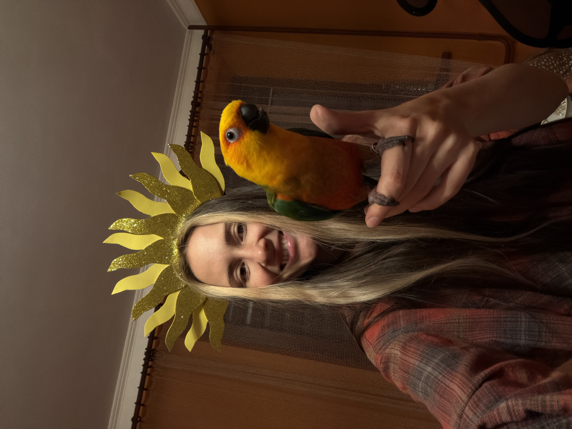 A young woman with long blonde hair, wearing a sunflower-themed headband, smiles warmly at the camera. She is holding a vibrant yellow, orange, and green parrot, likely a Sun Conure, perched on her left hand near her head. The woman wears a plaid shirt with dominant red and dark tones, and dark, textured rings are visible on her fingers. The image appears to be a selfie taken indoors in Khimki, Russia. In the background, a light-colored wall is visible on the left, while a window with a sheer, textured curtain or blind and a wooden rod can be seen on the right. The indoor lighting is dim. The woman's cheerful expression and the interaction with her pet parrot create a personal and happy atmosphere. The photo is rotated 90 degrees clockwise. No text is visible in the image.