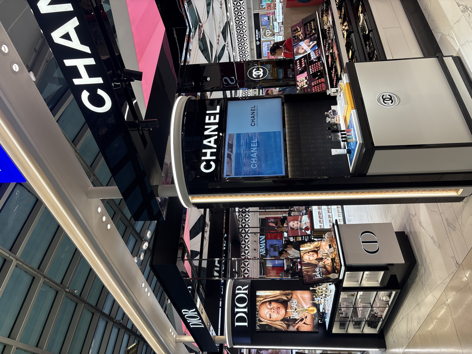 The image shows a high-end cosmetics store, likely in an airport duty-free shop given the bright lighting and polished floors. The store features prominent displays for Dior and Chanel, two luxury brands. The Dior section includes a large advertisement featuring a woman and bottles of perfume, as well as shelves stocked with skincare and fragrance products. The Chanel section displays various makeup products, including eyeshadow palettes and lipsticks, and features a screen showing a Chanel advertisement with the tagline "DIVE INTO SUMMER BEAUTY." Several staff members are visible behind the counters, and customers are browsing the merchandise in the background. The overall environment is clean, modern, and well-lit, with a focus on product presentation.
