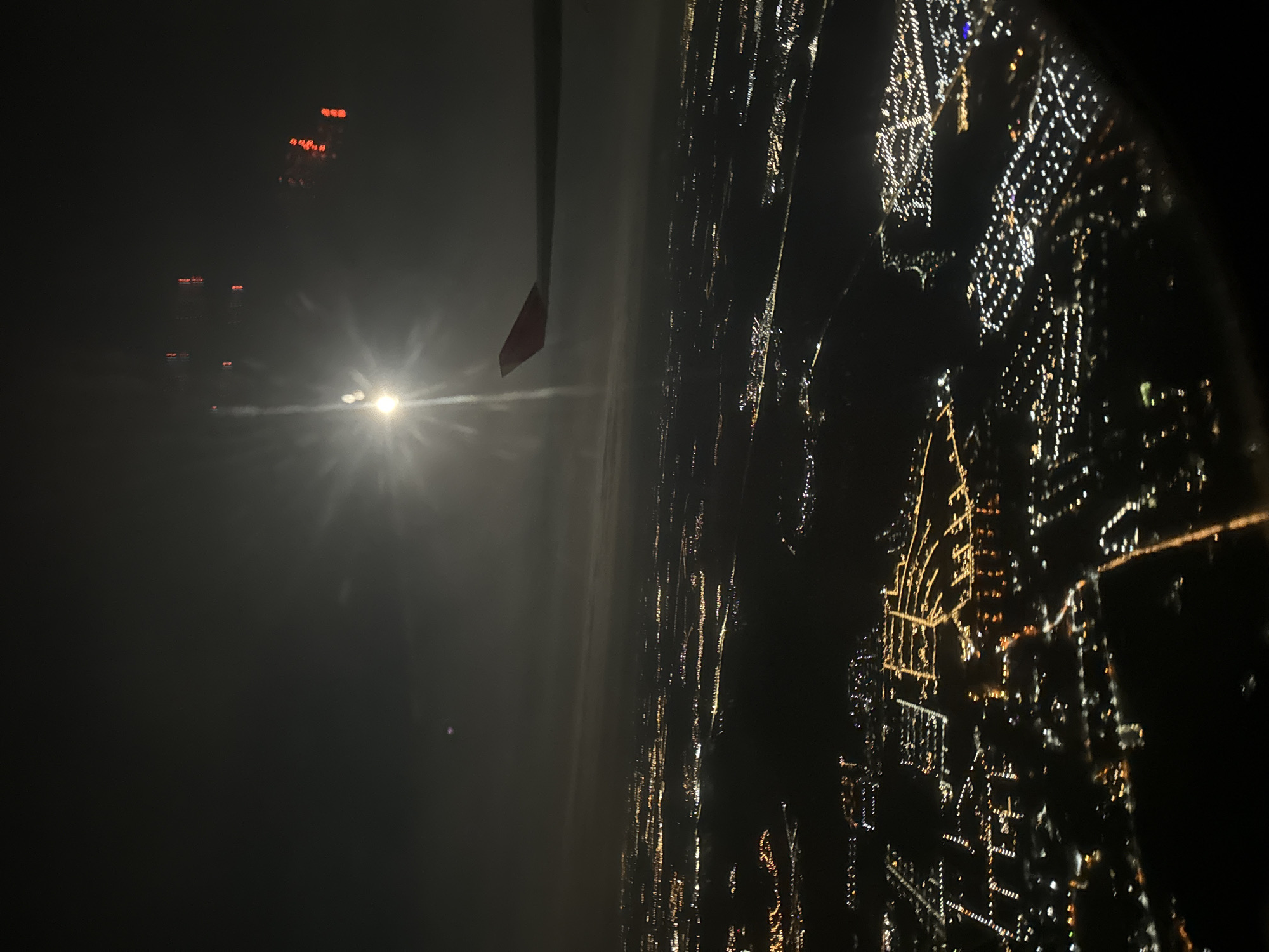 An aerial nighttime view from an airplane window shows the city lights of Troitsk, Russia. The wing of the plane is partially visible. A bright light source is to the left of the wing, possibly the moon or sun. City lights form a grid-like pattern below, interspersed with darker areas.