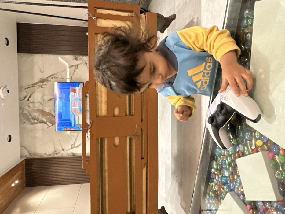 The image displays a young child interacting with a video game controller in an indoor setting. The image is oriented vertically (rotated 90 degrees clockwise from its natural upright position).
The central focus is a young child, likely a toddler or preschooler, with dark, somewhat messy hair. The child is wearing a two-toned long-sleeved shirt: the main body is light blue, and the sleeves and collar are yellow. The brand name "adidas" is prominently displayed in yellow text on the blue portion of the shirt. The child's face is looking downwards, with eyes appearing to be closed or cast down intently. Their left hand is resting on a white PlayStation 5 DualSense controller, which is placed on a clear glass table. The child's right hand is slightly clenched.
The glass table beneath the controller contains a decorative layer of numerous small, polished, and colorful stones or marbles. These stones are mostly iridescent or translucent, with dominant hues of blue, green, and pink, creating a vibrant base beneath the transparent surface.
In the background, behind the child and the table, there is a piece of light brown wooden furniture, possibly a cabinet or part of a bed frame, featuring vertical slat details. Beyond this furniture, a wall with a prominent white and gray marble pattern is visible. Mounted on this marble wall is a flat-screen television, which is actively displaying content. The screen shows bright blue and purplish colors, suggesting a video game or animated program.
Below the marble wall, there's a horizontal band of dark brown paneling, possibly made of wood or a similar material with a textured, slatted appearance. To the far left side of the image, a white wall is visible, on which a white device resembling a small security camera or sensor is mounted. Further down on this white wall, two small, circular, dark fixtures (possibly light sources or outlets) are visible. The floor appears to be a light-colored, possibly tiled, surface.
The lighting suggests an indoor setting, likely daylight or bright artificial light. The overall scene conveys a moment of focused engagement as the child interacts with the gaming controller.