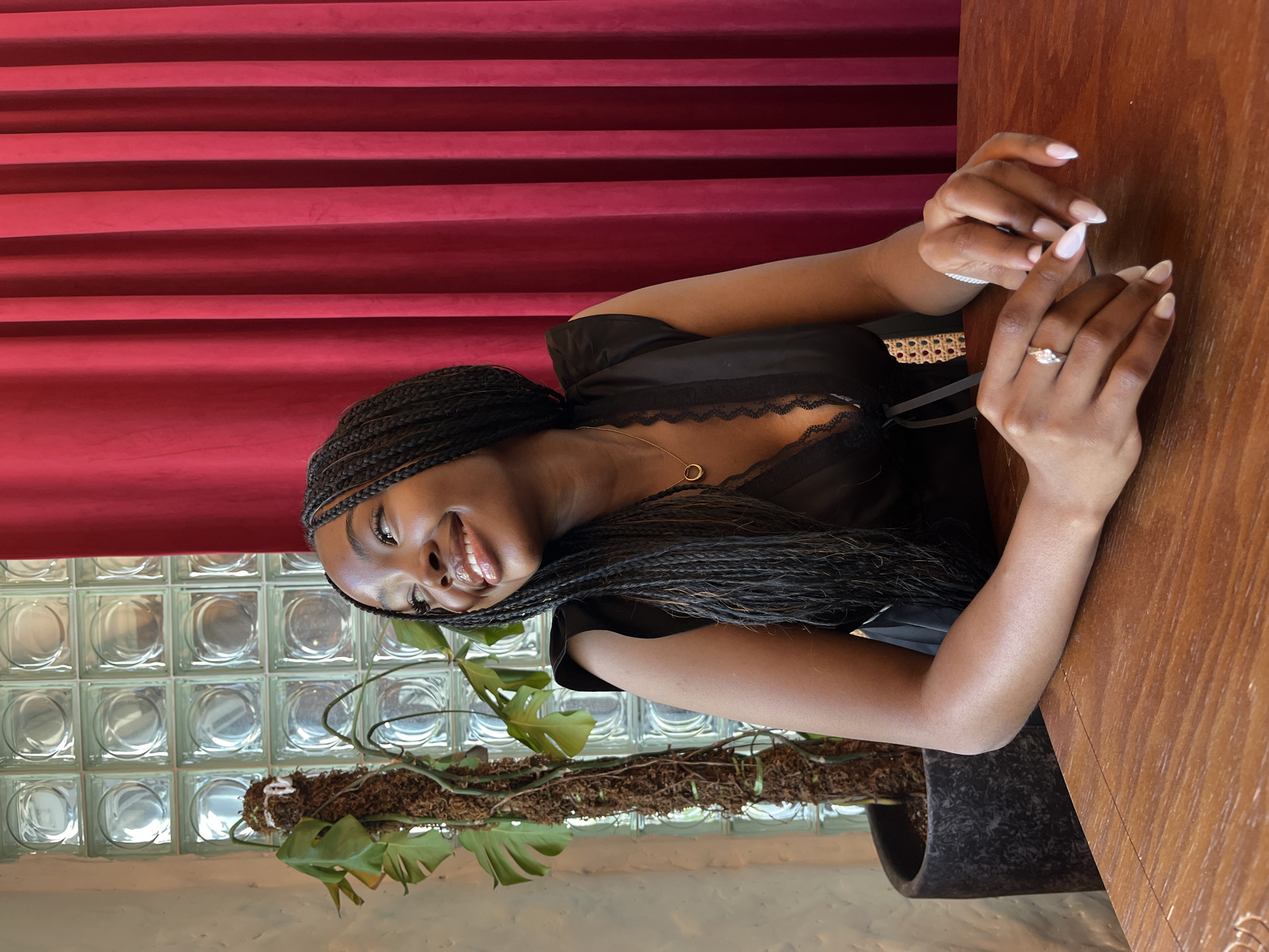 The image shows a young Black woman sitting at a wooden table in an indoor setting. She is wearing a black dress and has her hair styled in long braids. A ring with a stone is visible on her left hand, and her nails are painted a light color. In the background, there is a wall with glass blocks, a potted plant with large leaves, and a red velvet curtain. The lighting suggests an indoor environment, possibly a cafe or restaurant. The image does not provide any information about the location being in Kazan, Russia.