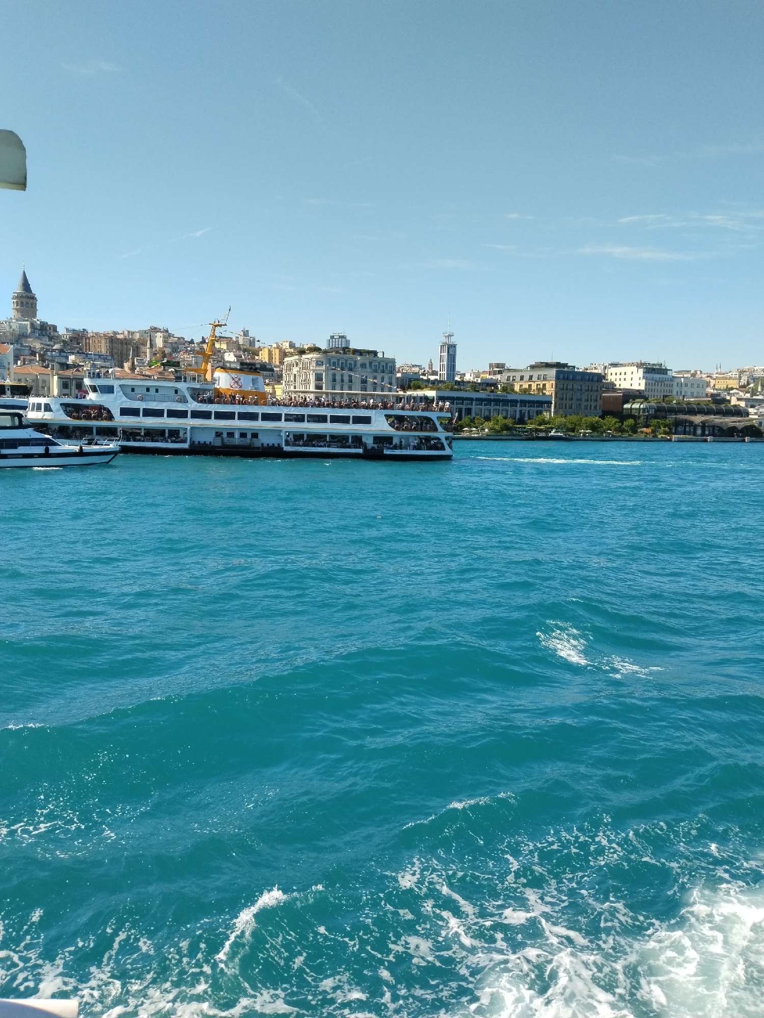 The image depicts a bright, clear day on the water in Sarıyer, Turkey. The sea is a vibrant turquoise, with visible waves and white foam from boat wake in the foreground. In the mid-ground, there are two large, white passenger ferries, one appearing to be docked or moving slowly, while the other is closer to the shore. The ferries are filled with people, mostly visible as dark shapes.
The background shows a city skyline with a mix of buildings. The Galata Tower is visible on the left, marking the location as Istanbul. Other buildings vary in height and architecture, indicating a dense urban environment. The sky is a clear blue with minimal cloud cover. A piece of white material is visible in the top left corner.