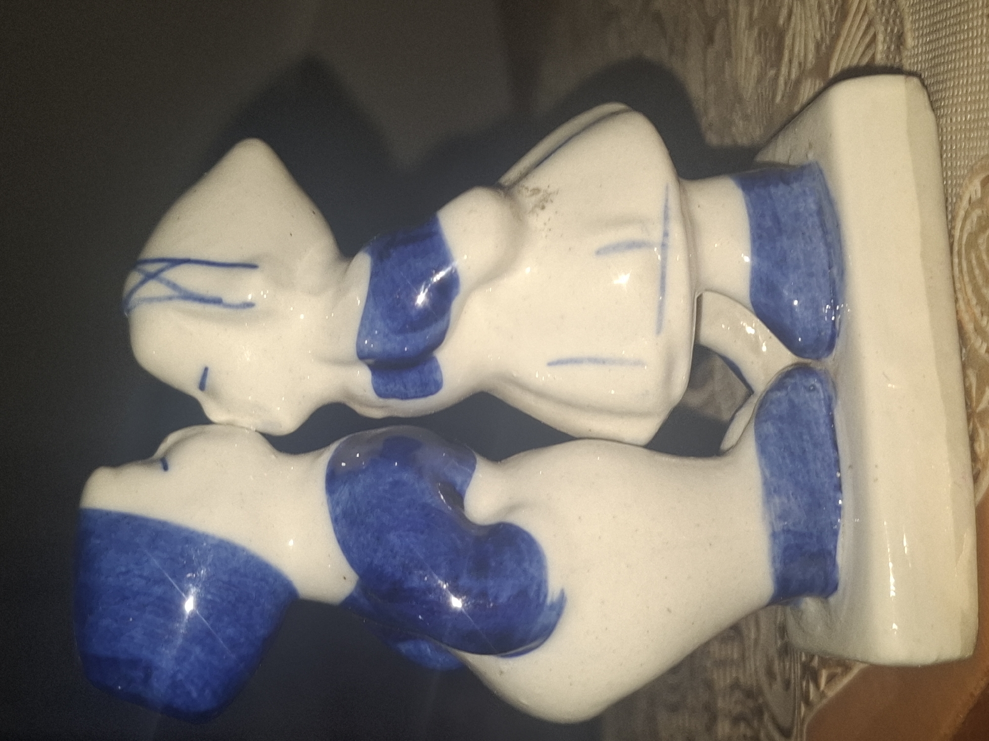 A ceramic figurine of two children kissing is displayed on a textured surface. The children are depicted with white skin and blue clothing details, including their hats, shirts, and shoes. The figurine stands on a white rectangular base. The background is dark and out of focus. The figurine is positioned in what appears to be an indoor setting. There is no discernible indication of the city of Almaty, Kazakhstan, or any specific urban environment.