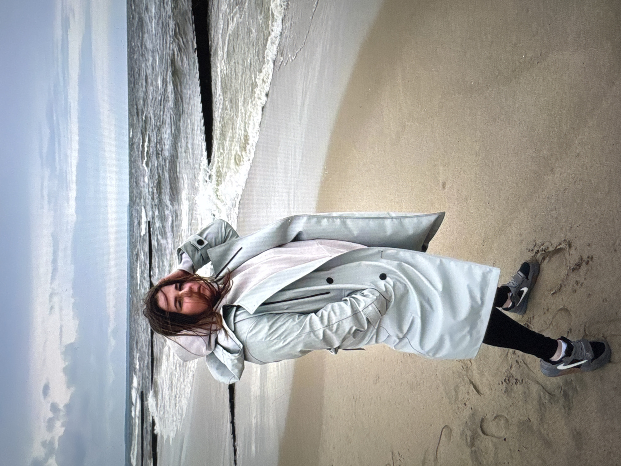 The image depicts a woman lying on a sandy beach next to the ocean. She is wearing black leggings, grey sneakers, a light pink shirt, and a light blue trench coat. The woman is lying with her head resting on her arm. The background includes a cloudy sky, the ocean with visible waves, and wooden posts jutting out of the water. The sand appears light tan in color. The location is likely near Новинки, Russia, given the presence of beaches on the coastline there.