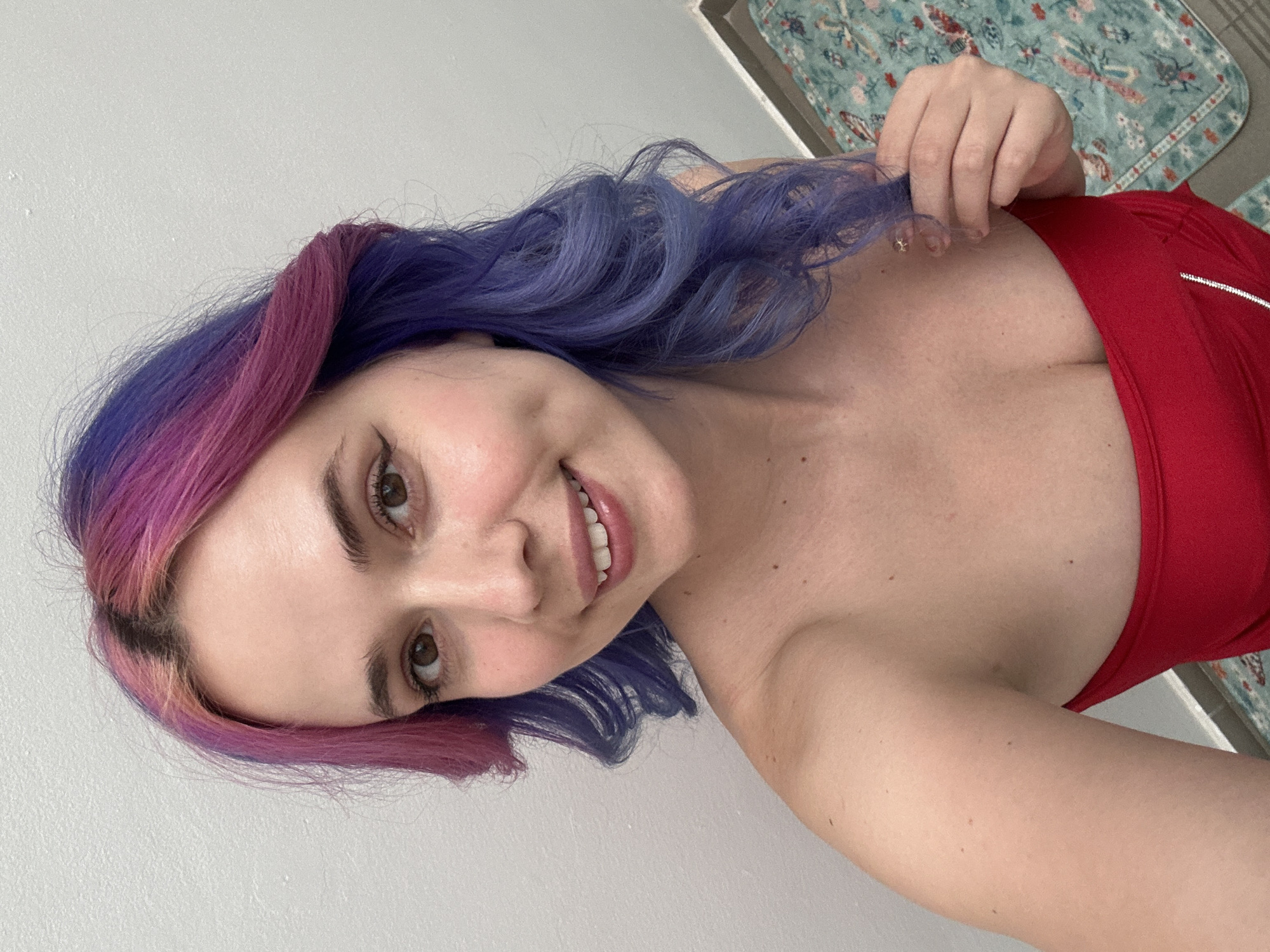 A close-up selfie captures a young woman with vibrant purple and pink ombre hair, smiling warmly at the camera. Her head is tilted, and her long, wavy hair cascades around her face and shoulders. With her right hand, she gently pulls on a strand of her purple hair, which shows off its distinct color. Her brown eyes are wide, and her lips are slightly parted in a cheerful smile, revealing her teeth. She appears to be wearing a red top or dress with a low neckline, and a white, possibly reflective, stripe or zipper detail is visible on the garment over her right shoulder.
The background consists primarily of a plain, light-colored wall, suggesting an indoor setting. In the upper right corner, a segment of a patterned mat or rug with teal, red, and beige floral or foliage designs can be seen, adding a touch of color to the otherwise neutral background. The lighting is soft and even, highlighting her clear complexion. The overall impression is one of a friendly and confident individual. The photograph is taken in what is understood to be the City of Zagreb, Croatia.