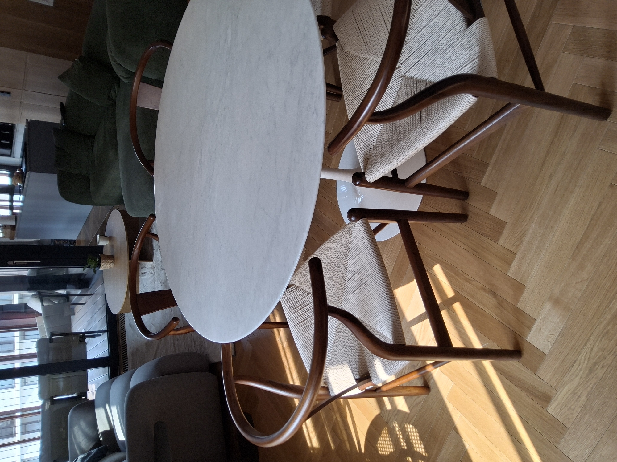 The image depicts a modern dining area with a round, white marble-top table supported by a white pedestal base. Four wooden chairs with woven seats are arranged around the table. The chairs have curved backs and wooden frames. The flooring is a light-colored wood laid in a herringbone pattern. Sunlight streams in from the left, casting diagonal shadows across the floor and the chairs. In the background, to the left, a portion of a living room is visible, featuring a light-colored sofa and a large window with vertical blinds. Further back, to the right, a dark green sectional sofa is visible. The scene appears to be indoors, likely in a residential apartment or a commercial space in Warsaw, Poland. No people are present in the image.