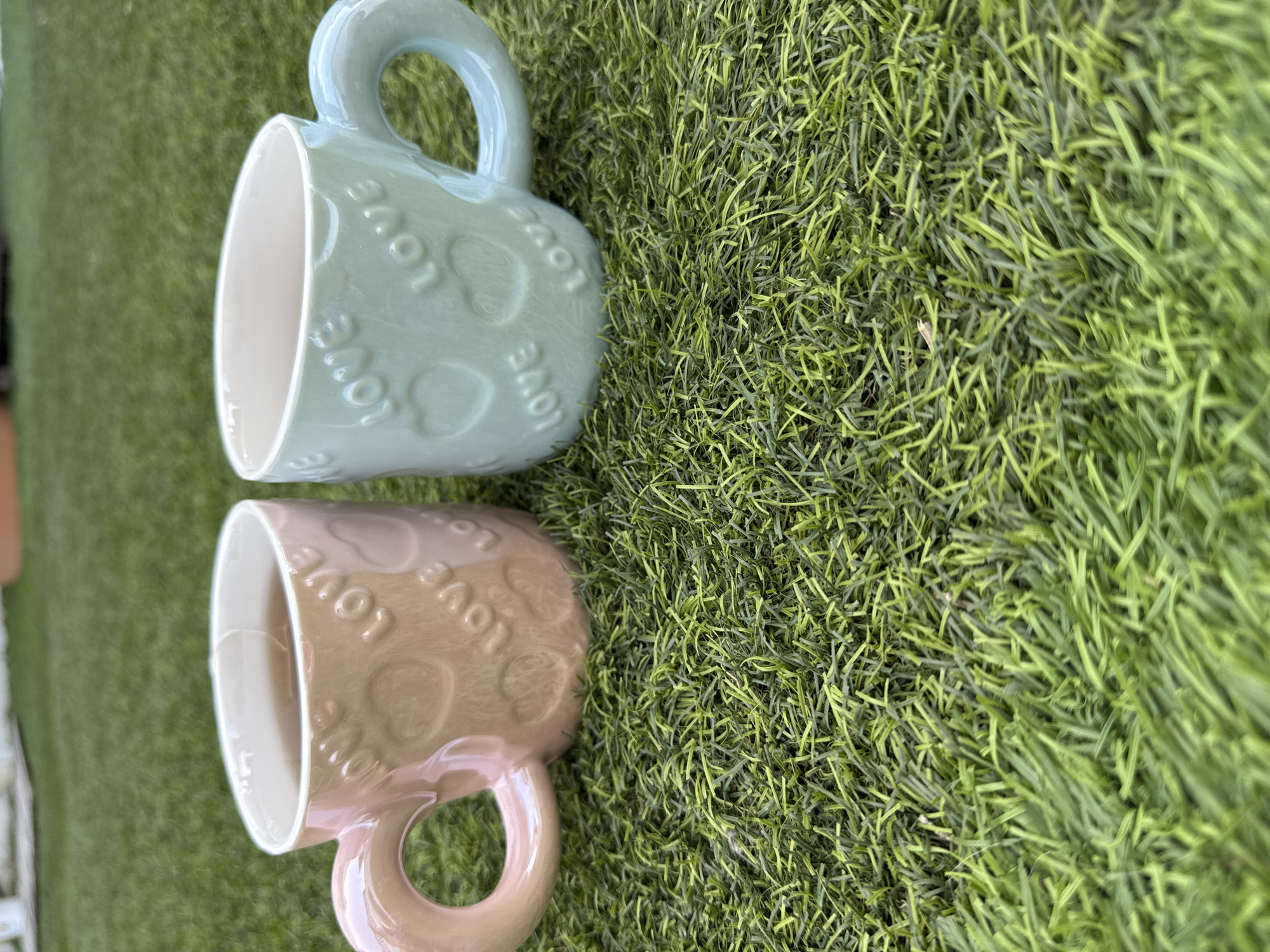 Two ceramic mugs are placed on artificial grass. The mug on the left is pink and contains a dark liquid. The mug on the right is light blue and is empty. Both mugs have raised hearts and the word "LOVE" embossed on their sides, as well as heart-shaped handles. The background consists of more artificial grass. The scene appears to be outdoors, possibly in a garden or yard. There is no indication of the location being in Vallada, Spain.