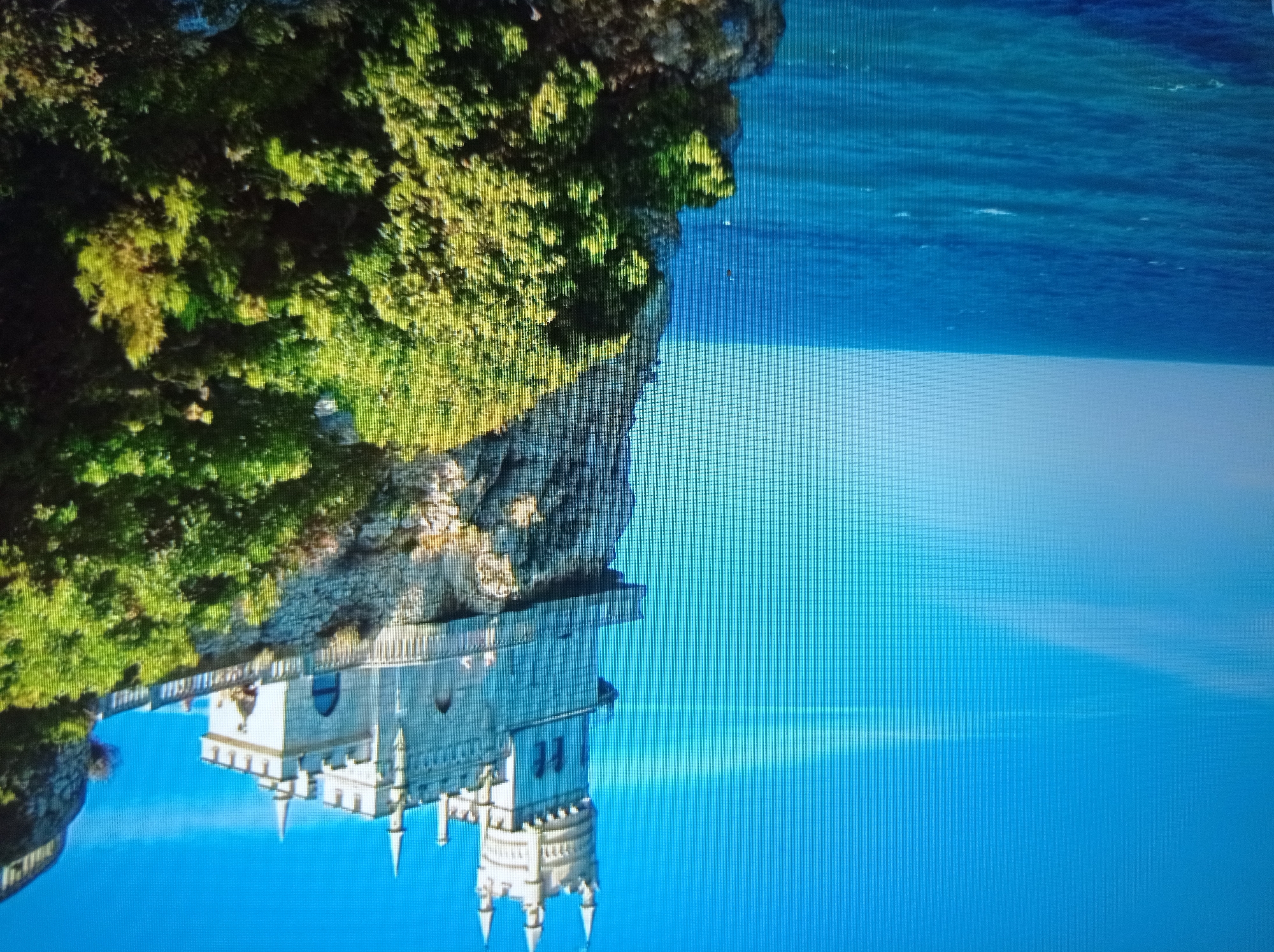 The image depicts a castle perched precariously on a cliff overlooking a body of water. The castle is made of light-colored stone and features multiple turrets and battlements, giving it a Gothic or medieval appearance. A balcony or walkway extends from the castle, offering a view of the sea. The cliff face is rugged and covered in lush green vegetation, including trees and bushes. The sea is a deep blue, with gentle waves visible. The sky is a bright, clear blue, with a few wispy clouds. The overall impression is one of a dramatic and picturesque landscape.
This image is not located in the city of Mukachevo, Ukraine. The castle shown is the Swallow's Nest Castle, located in Crimea.