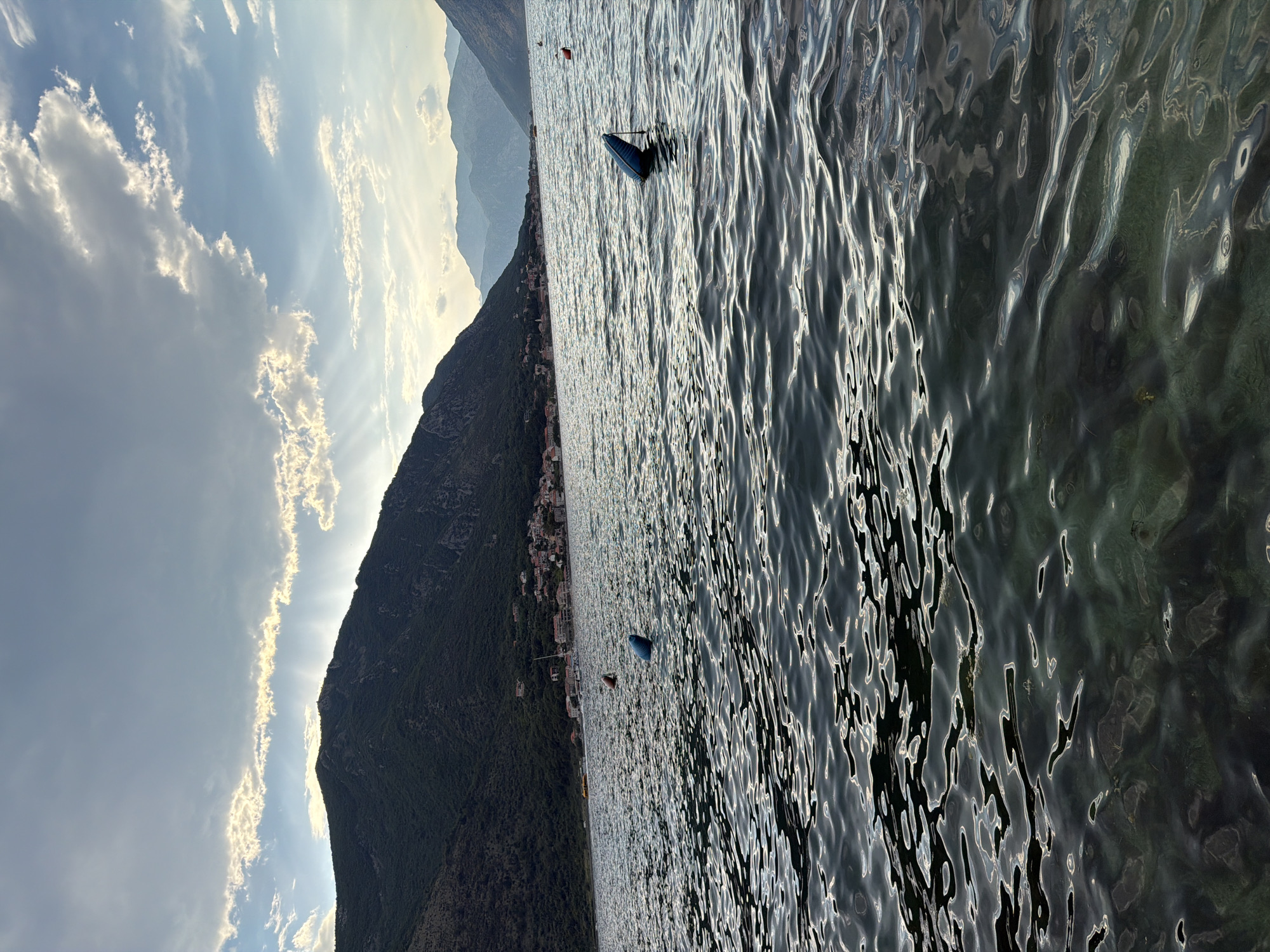 The image depicts a tranquil scene in Dobrota, Montenegro, characterized by the Bay of Kotor and its surrounding mountains under a dramatic sky. The foreground is dominated by the rippling surface of the bay, with sunlight glinting off the water, revealing hints of submerged rocks and vegetation below. Several blue buoys are visible in the water, marking an area of the bay. In the mid-ground, a picturesque coastal town with red-tiled roofs is nestled at the base of a lush, green mountainside. The mountains rise steeply, covered in dense vegetation, and extend into the background. The sky above is a canvas of light and shadow, with scattered clouds allowing beams of sunlight to pierce through, creating a picturesque, almost ethereal effect. The overall impression is one of natural beauty and serene coastal living.