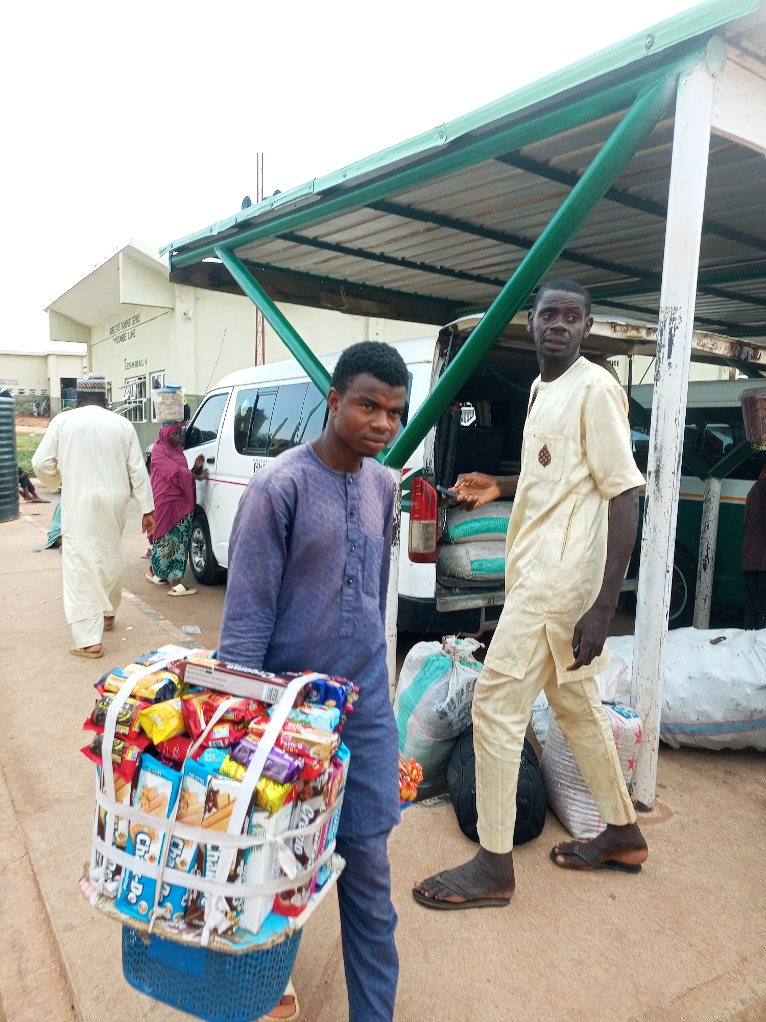 The image depicts an outdoor scene at a transport hub, likely a bus station or a similar terminal, in Gombe, Nigeria. Two men are the central figures. The man in the foreground, to the left, is wearing a blue buba and is carrying a large basket filled with assorted snacks and confectionery. The basket is tightly wrapped with white tape to secure its contents. To his right, a man in a cream-colored agbada stands facing forward, holding a mobile phone in his left hand. Behind them, a white commercial bus is parked under a green metal canopy. In the background, a building with "TERMINAL 4" and "GOMBE LINE" signage is visible. Other individuals are present in the background, including a woman in a maroon headscarf and a man in a white agbada walking away from the camera. Large sacks are piled near the bus. The ground is paved with asphalt. The overall impression is that of a busy public transportation area where vendors are actively engaged in commerce.