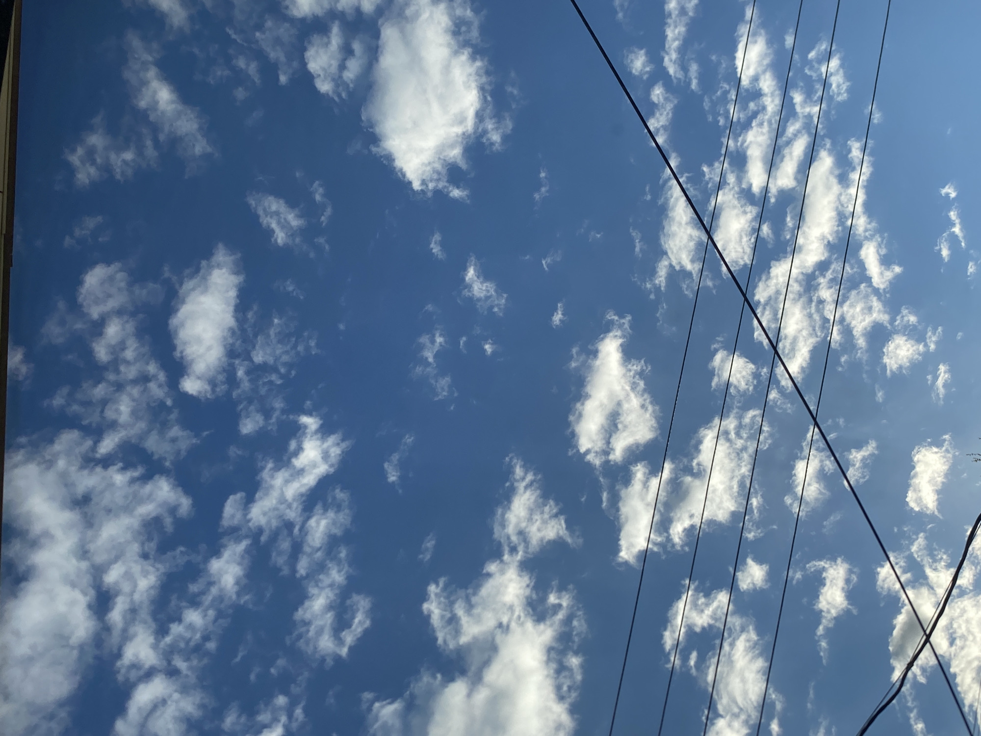 The image shows a view of the sky, possibly in Фоки, Russia, though this cannot be confirmed from the image itself. The sky is blue with scattered white clouds. Several black power lines or cables are visible running diagonally across the frame, from the upper right to the lower left, partially obstructing the view of the sky.