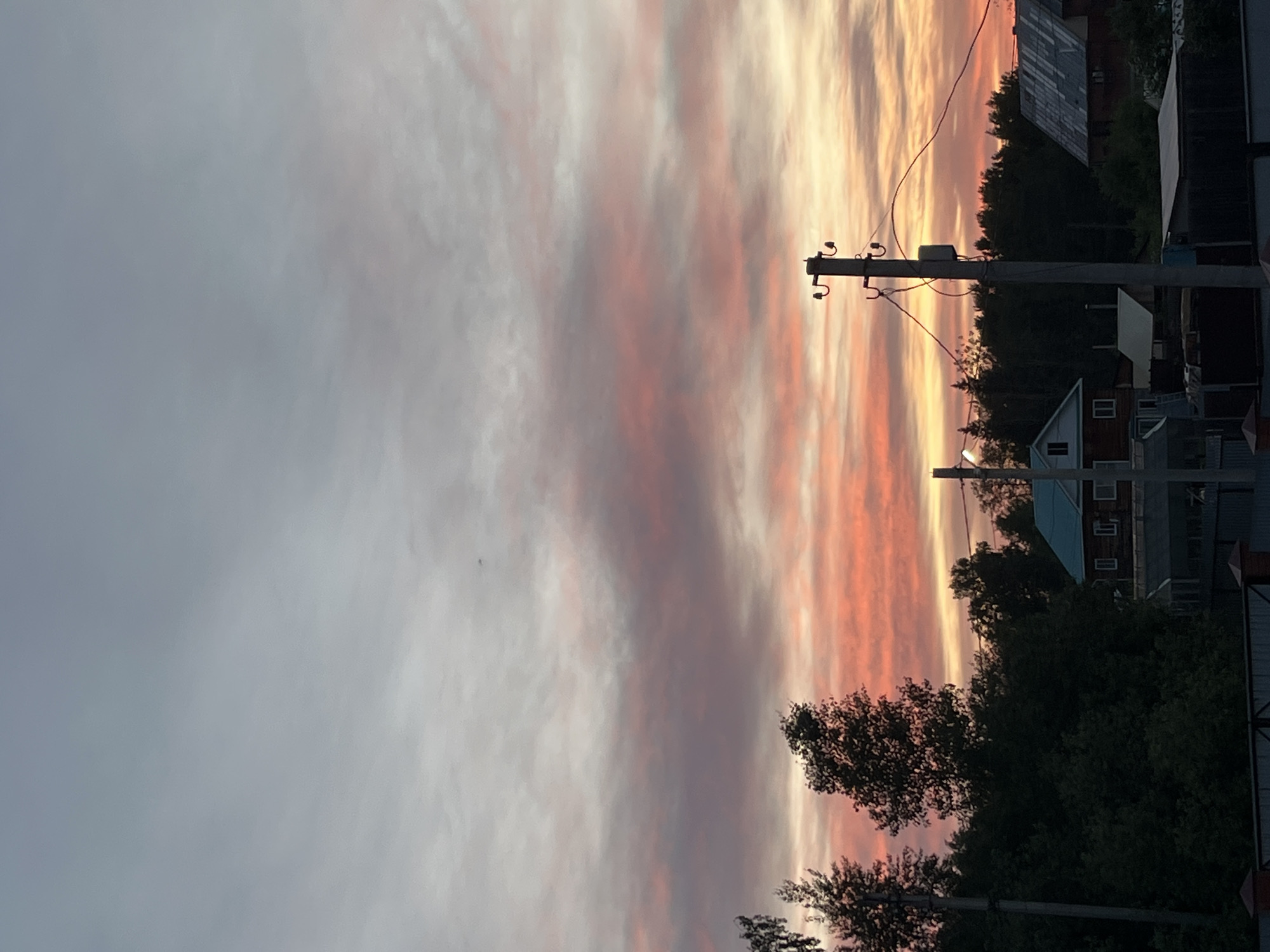 The image shows a colorful sunset sky with pink, orange, and yellow streaks. In the foreground, there are trees and utility poles with wires running across the sky. Further in the background, there are houses and buildings. The image appears to be taken in a residential area. While the image resembles scenes in Chaikovsky, Russia, it is impossible to confirm the location without additional information.