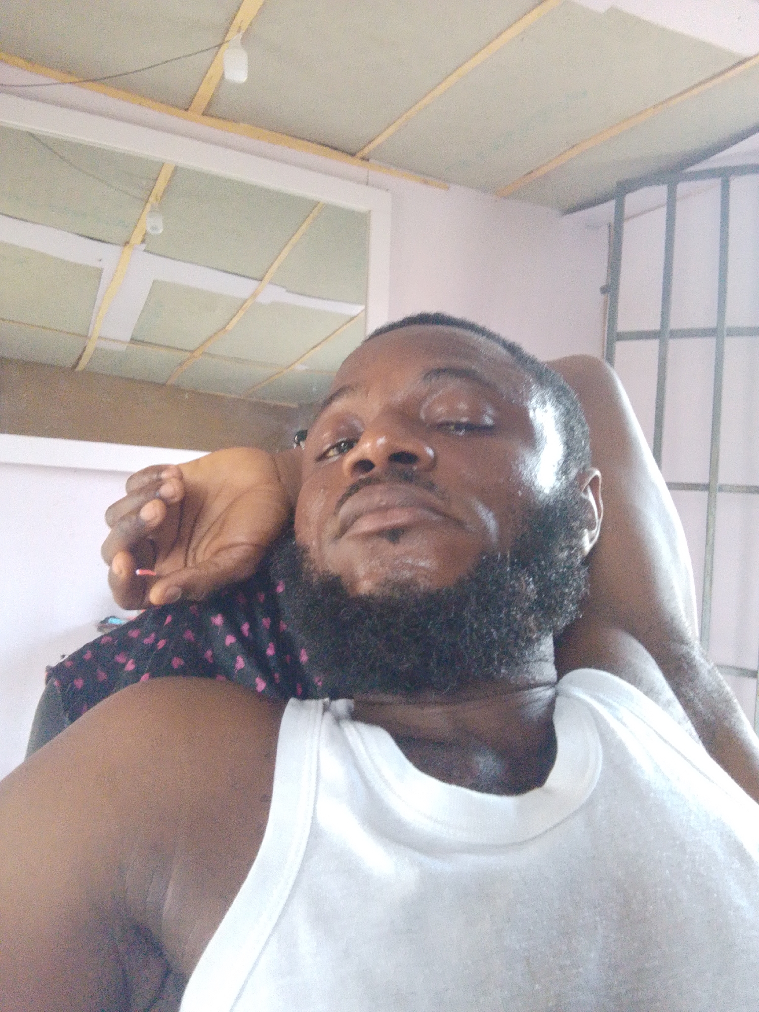 A close-up, selfie-style photograph features a Black man with a beard wearing a white tank top. His right arm is raised and positioned behind his head, while his left hand rests on his chest. He appears to be in an indoor setting, possibly a residential space, based on the visible ceiling with exposed wooden beams and drywall panels, as well as a metal grid structure in the background. The location in the city of Lokoja, Nigeria cannot be determined from the image.