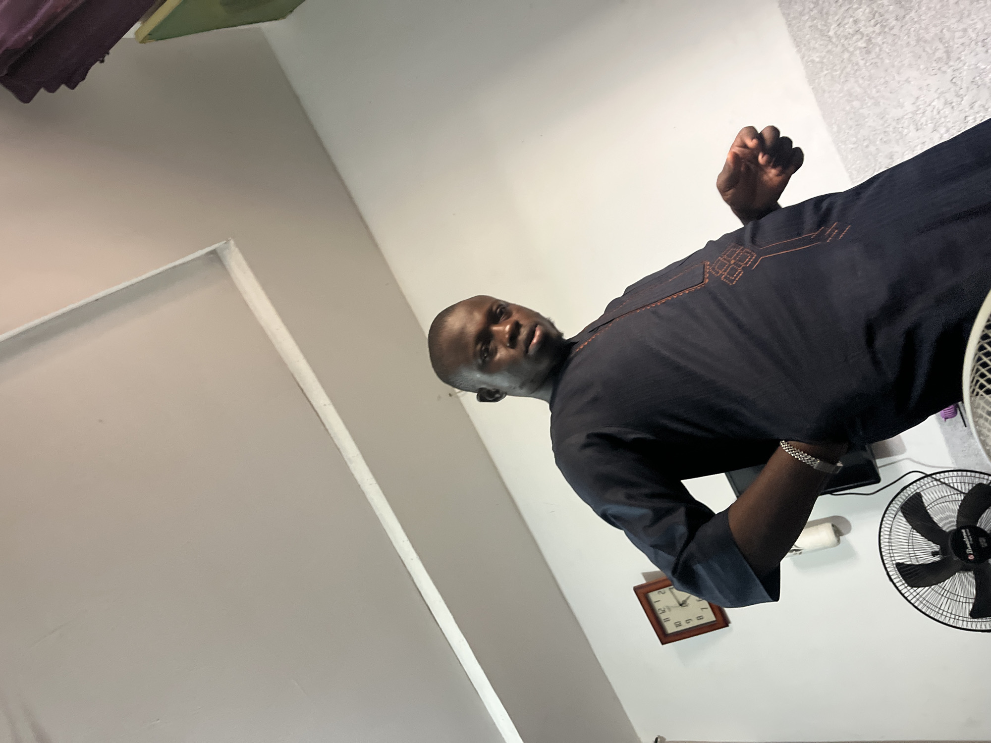 A man is indoors in what appears to be a residential setting. He is wearing a dark garment with orange embroidery, standing near a white wall with a clock and a fan visible in the background. The location is suspected to be in the city of Abuja, Nigeria, based on the cultural clothing.
