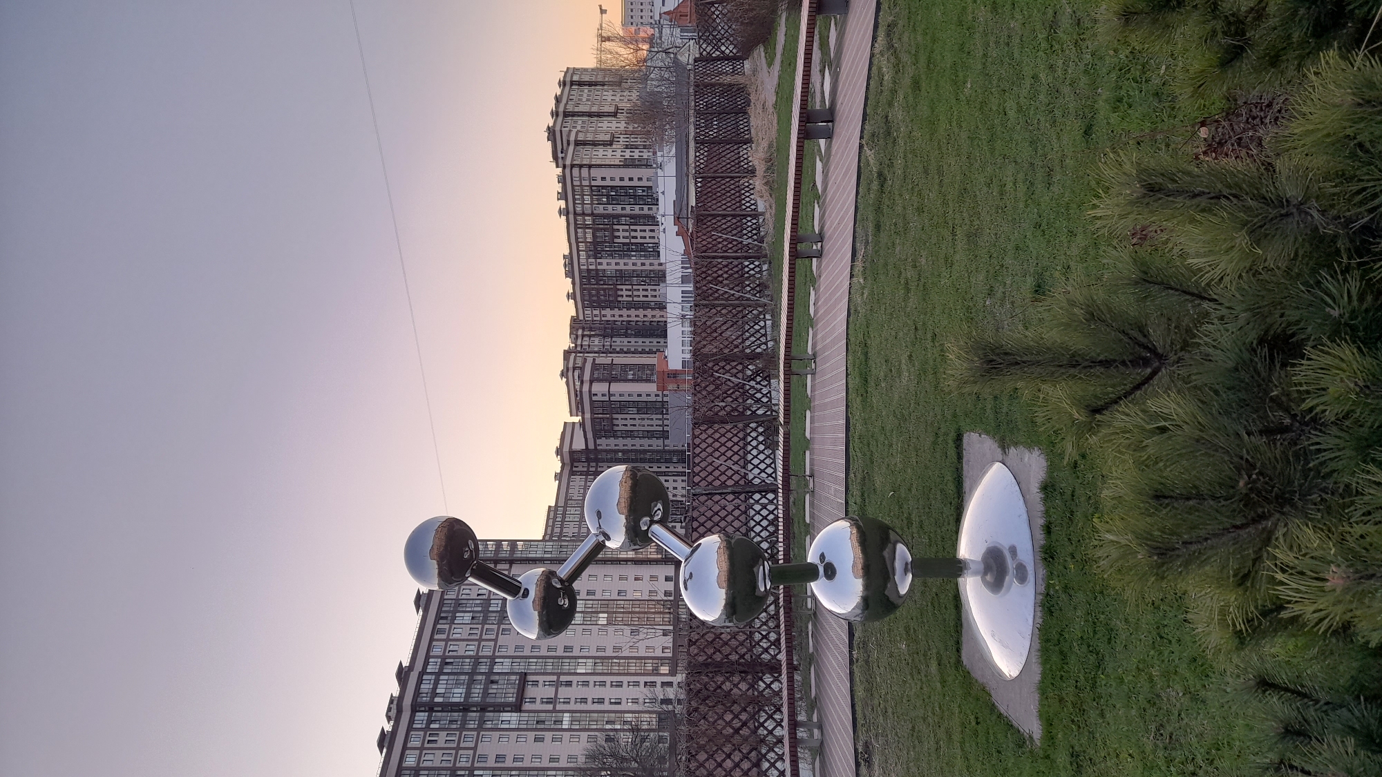 The image depicts a park scene in Saint Petersburg, Russia, at sunset. The park features a modern art installation of interconnected metallic spheres, resembling a molecular structure, situated near a walking path. Adjacent to the path is a low, decorative fence, separating the path from a grassy area. In the background are multi-story residential buildings, partially reflecting the sunset hues. A pine tree stands on the right side of the image, partially obscuring the view.