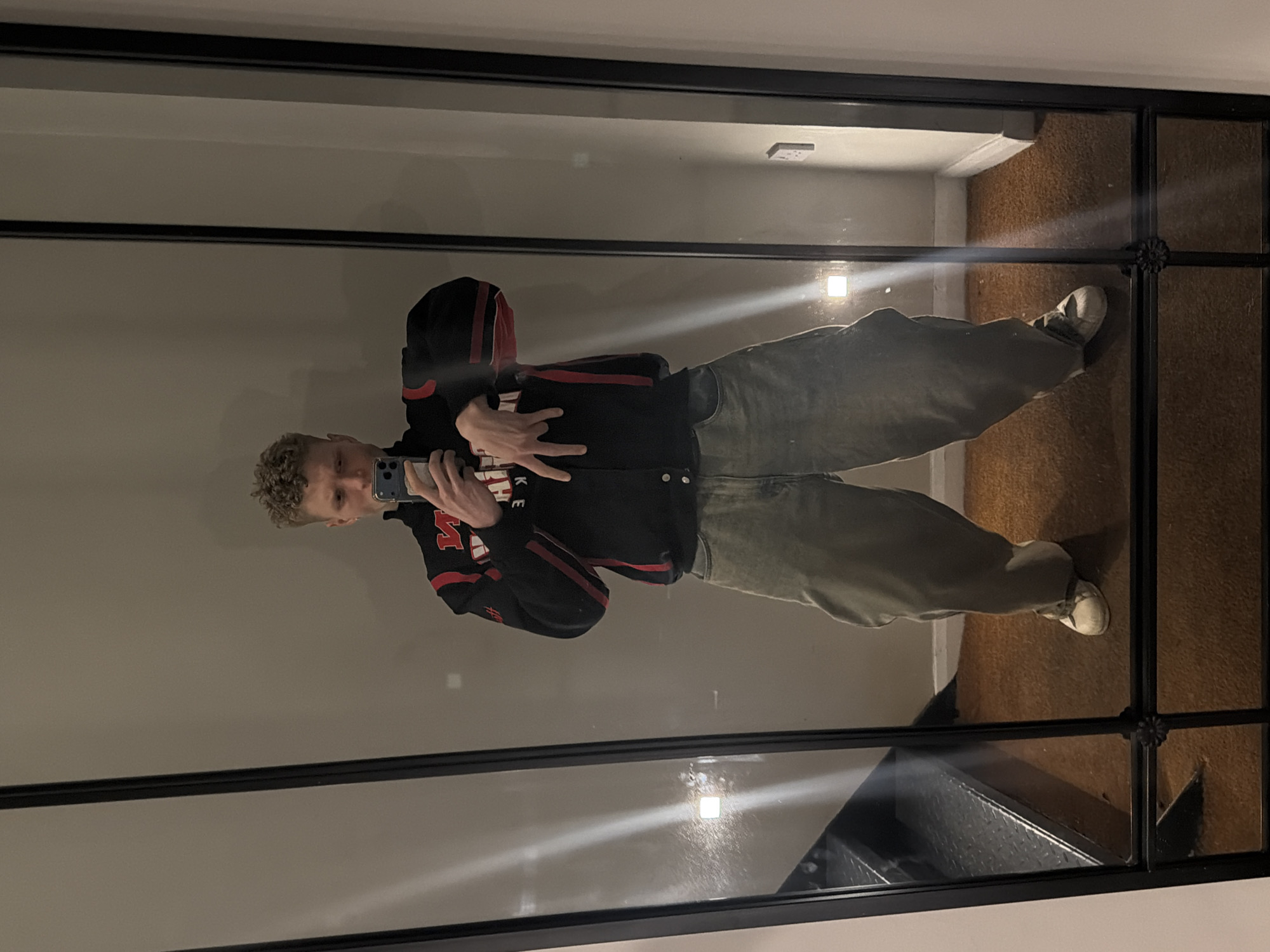 A male individual is depicted in a full-body reflection within a large, black-framed mirror. He has short, curly, light-colored hair and an earring in his visible left ear. He is holding a dark-colored smartphone in his right hand, pointed towards the mirror, seemingly capturing the image. His left hand is raised, performing a "rock on" gesture with the index and pinky fingers extended. He wears a black varsity-style jacket with red and white accents, featuring white buttons down the front. Inverted white lettering is visible on the left chest area, possibly "MNL" or similar. He is also wearing light-colored, baggy jeans or trousers of a faded green-grey hue and white sneakers. The environment reflected in the mirror consists of plain, light-colored walls and a dark orange-brown carpeted floor. Two square, recessed light fixtures are visible on the side walls, along with a white electrical outlet or switch on the right wall. Through the bottom left pane of the mirror, a section of dark, industrial-style stairs with a textured surface is visible. No specific visual cues are present within the image to definitively identify the location as the city of London, United Kingdom.