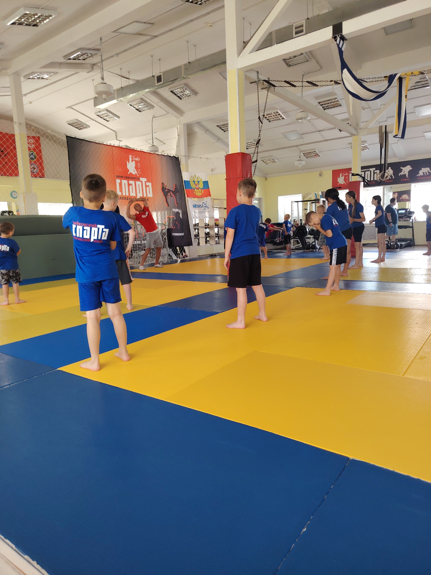 A group of children and a few adults are participating in a judo class in an indoor gymnasium. The gym has a blue and yellow mat floor, and the walls are painted yellow. There are various pieces of sports equipment visible, including mats, weights, and a treadmill. In the background, there are banners and flags displayed. The children are wearing athletic clothing, and some are in the process of training, while others are observing. The overall atmosphere appears to be one of organized athletic activity. This scene likely takes place in Chaikovsky, Russia, a city known for its sports facilities and dedication to athletic development, particularly in sports like judo.