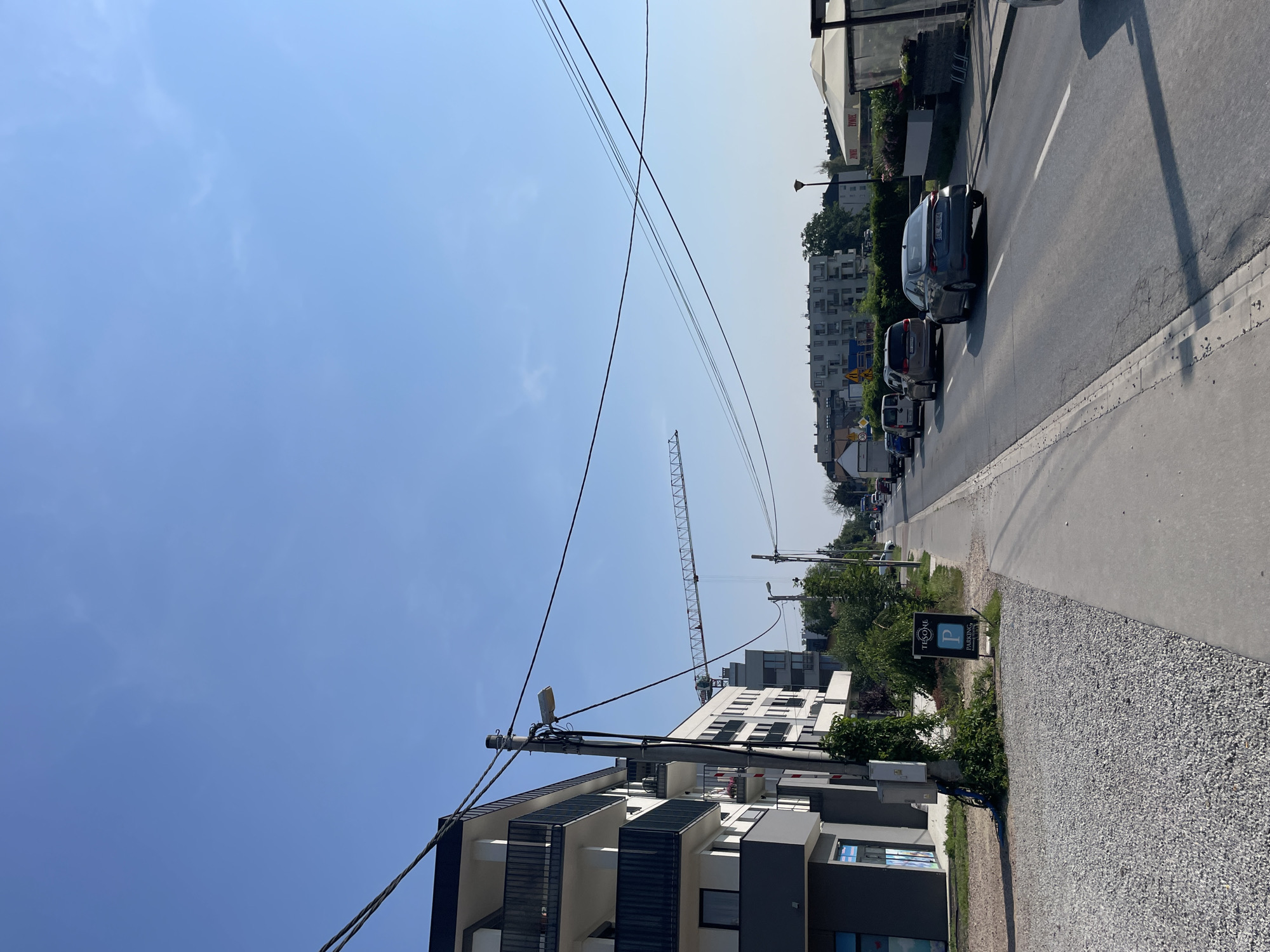 The image depicts a street scene with a modern apartment building on the left and a road with several cars on the right. The sky is clear blue. A construction crane is visible in the distance, suggesting ongoing development. A sidewalk runs alongside the street, with a parking sign on it. The apartment building has balconies with black railings and white facades. Trees and greenery are present along the sidewalk and in the background. The road appears to be a two-lane road with traffic moving in one direction. The overall impression is of a residential or mixed-use urban environment on a sunny day.