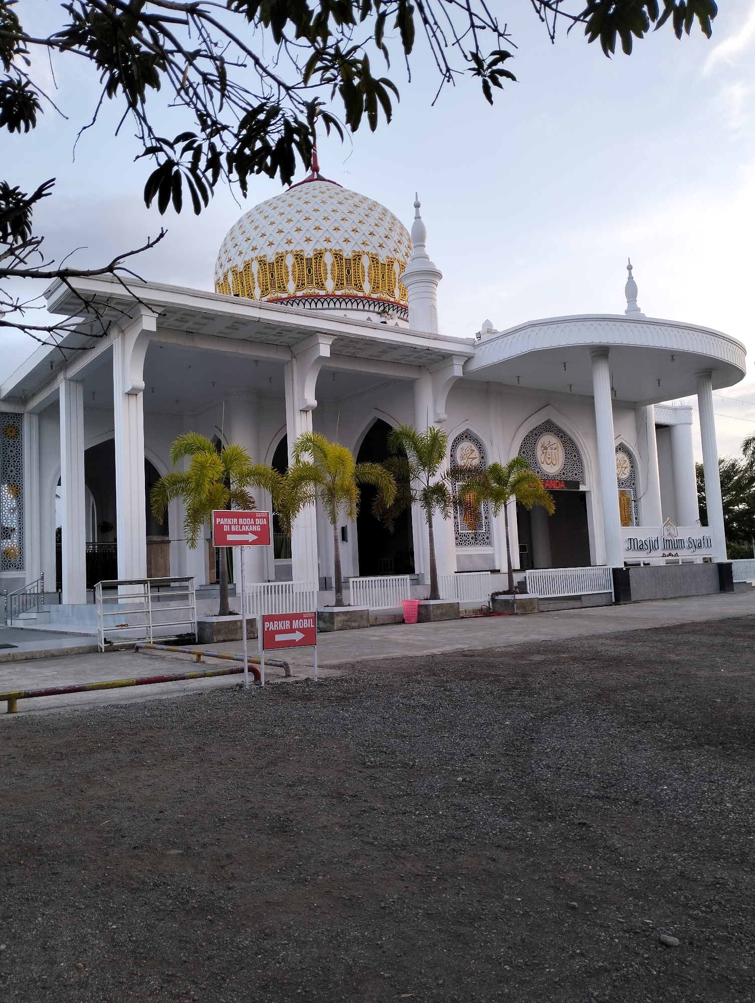 The image shows the exterior of a mosque, likely the Masjid Imam Syafi'i, located in Blangmetimu, Indonesia. The mosque is white with a prominent dome decorated with gold stars and intricate patterns. The building features large white columns supporting an arched roof structure. Several palm trees are visible in front of the mosque. Two red signs are posted, one indicating "PARKIR RODA DUA DI BELAKANG" (Motorcycle Parking in the Back) with an arrow pointing left, and another indicating "PARKIR MOBIL" (Car Parking) with an arrow pointing right. The ground in the foreground is covered with gravel. The sky is overcast. There are no people visible in the image.