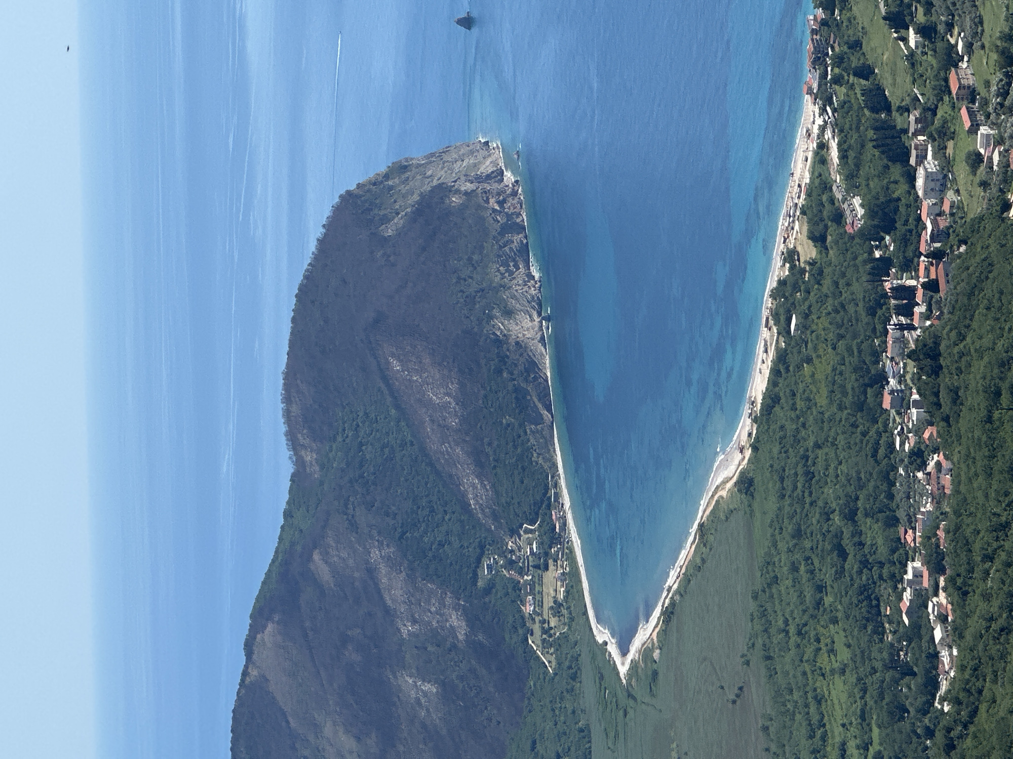 An aerial shot displays a coastline and mountainous terrain. The ocean is a gradient of blues, transitioning from lighter near the horizon to darker near the shore. A prominent headland extends into the sea, covered in vegetation and scorched patches. A narrow beach separates the water from the land. Buildings dot the coastline, interspersed with green trees. A structure in the water, perhaps a boat, is visible in the distance. The overall impression is one of a coastal landscape. This is Novoselje, Montenegro.