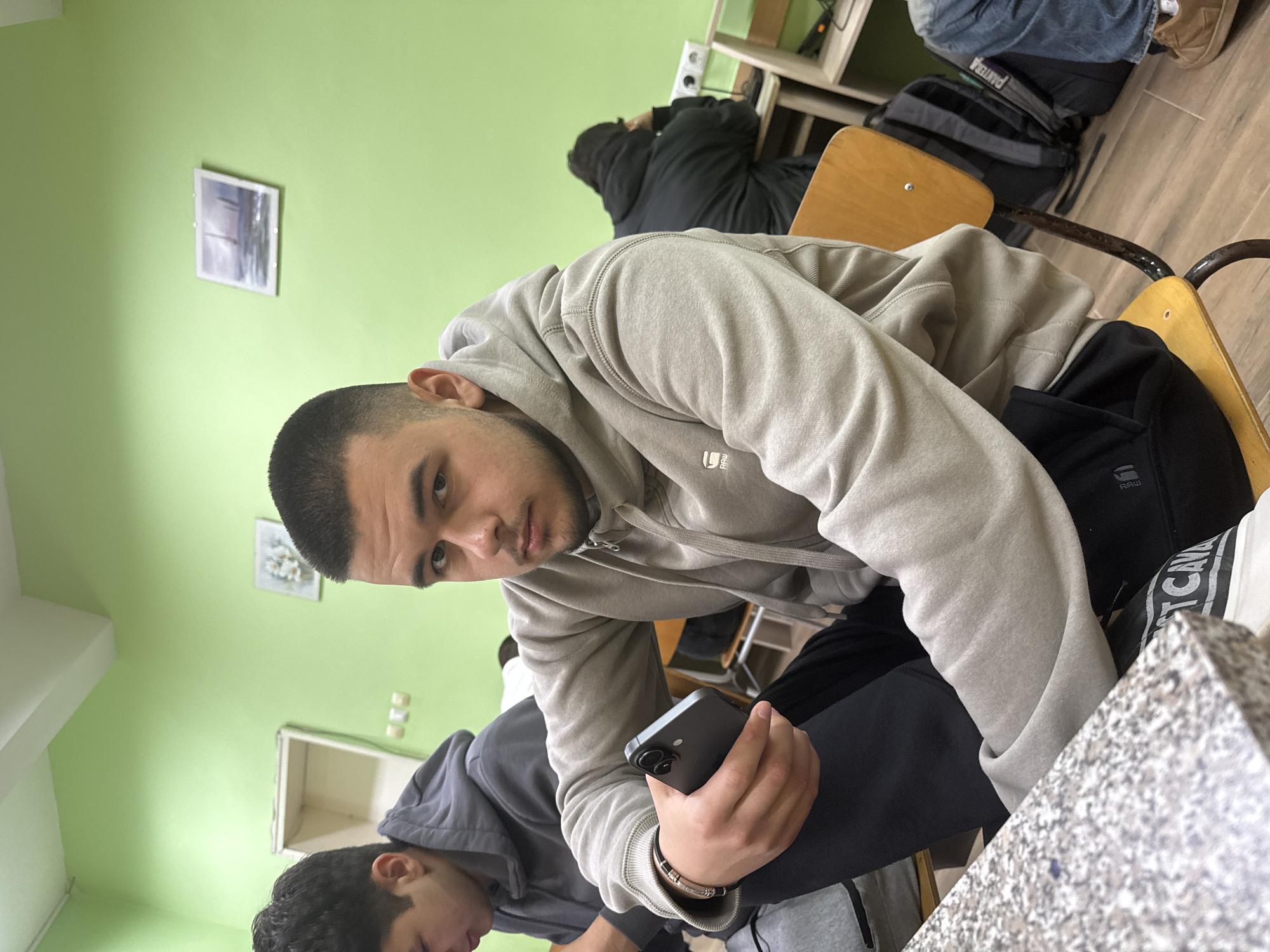 The image presents an indoor scene, likely a classroom or study area, characterized by light green walls and a wooden or wood-laminate floor. The photo is taken from a low, tilted angle, looking up towards the primary subject.
The main subject is a young man with a short haircut and a light beard, wearing a light gray hooded sweatshirt with a small, indistinct logo on the left sleeve (possibly "G-RAW") and dark pants. He is seated or leaning, looking directly at the camera with a neutral expression. He holds a dark-colored smartphone in his left hand and wears a bracelet on his left wrist.
In the mid-ground, another individual in a gray hoodie is partially visible, facing away from the camera. Further back, a person wearing a dark hoodie is seen from behind, possibly resting or engrossed in an activity. A black backpack with "PANTURA" visibly printed on it rests on a chair in the background.
The room is furnished with wooden desks and chairs. Two small, framed pictures are mounted on the light green wall, and an electrical outlet is also visible. A light-colored, possibly granite-patterned tabletop occupies the bottom right foreground. The overall ambiance suggests a casual moment within an educational or communal indoor space. The time of day is not discernible.