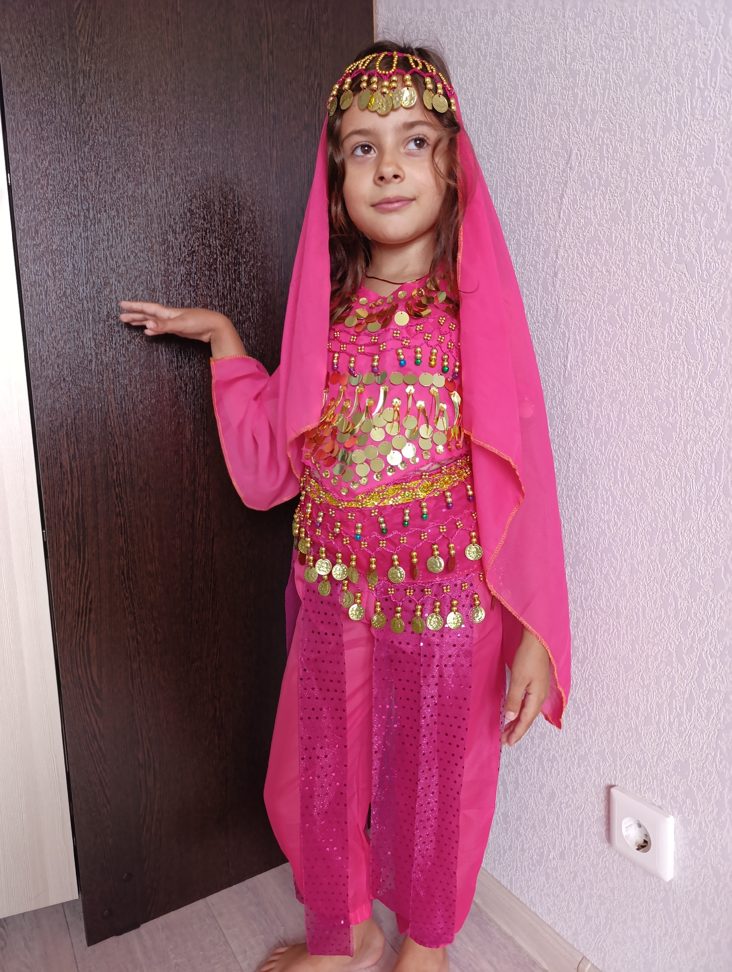 A young girl, appearing to be of European descent, stands in what appears to be a residential setting. She is dressed in a vibrant pink, sequined costume reminiscent of Middle Eastern attire, complete with a headpiece adorned with gold coins and beads. The costume includes a flowing veil, a decorated top, and baggy pants. Her left arm is extended, with her hand resting near a dark brown wooden door. She is looking slightly upwards and to her right, with a subtle smile. The background consists of a textured light pink wall with a white electrical outlet visible on the right side. The floor is light-colored laminate. There is no indication of the city of Mariupol, Ukraine, or any other specific location in this image.