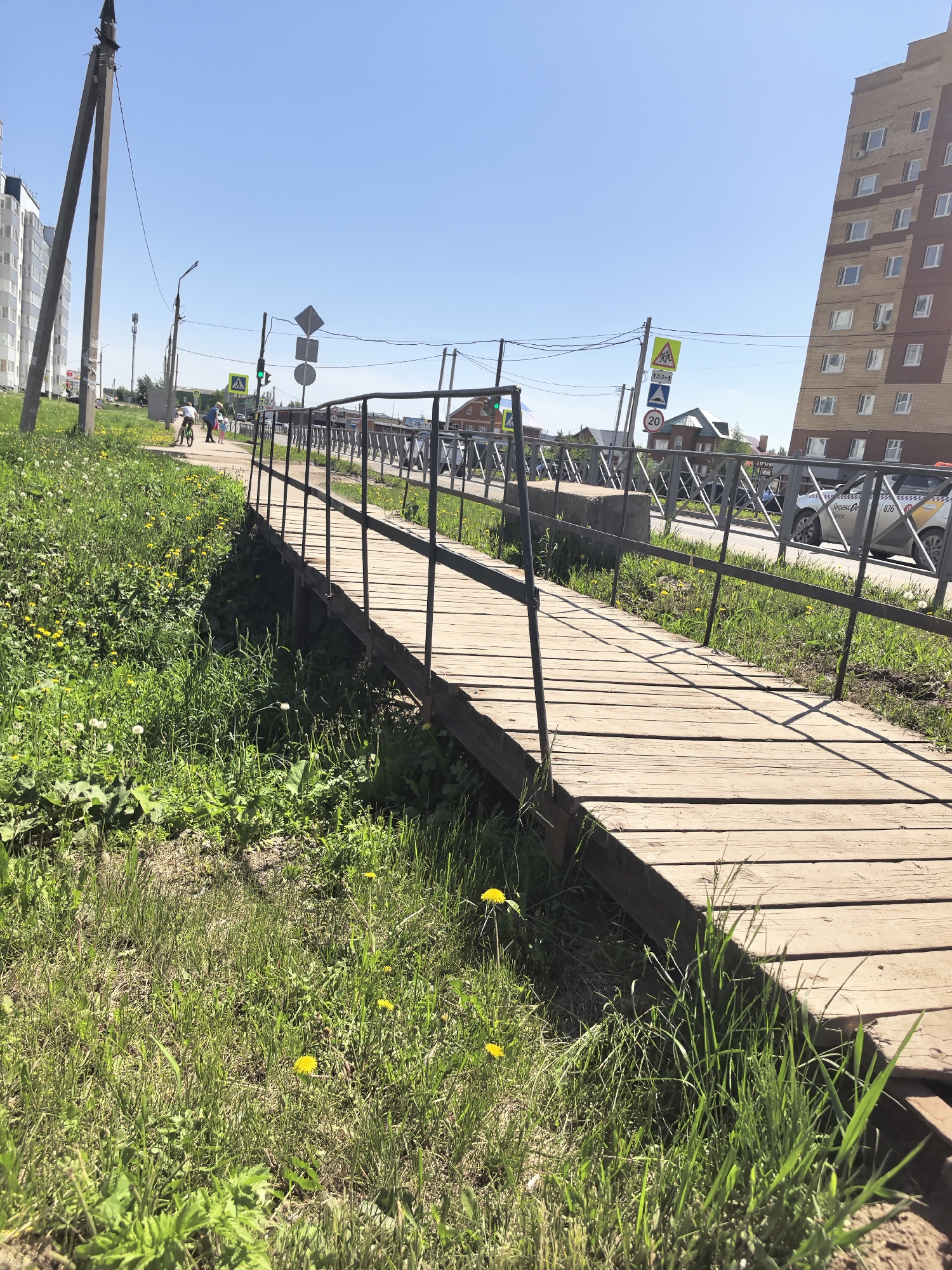 A wooden pedestrian bridge with metal railings spans a grassy area in the city of Фролы, Russia. The bridge deck is made of wooden planks. The railing consists of vertical metal bars connected to horizontal bars. The area surrounding the bridge is covered in green grass and dandelions. A group of three people, two adults and one child, are walking along a paved walkway adjacent to the bridge. Street signs and utility poles are visible along the walkway. An apartment building with windows is in the background. A car identified as a Яндекс Такси is on the street behind the bridge. The sky is clear blue.