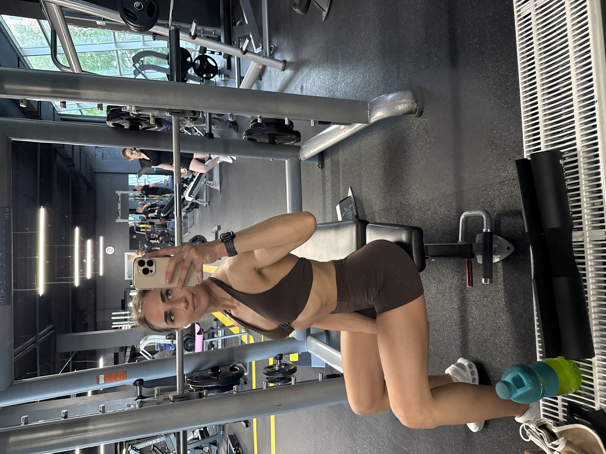 The image depicts a young adult female taking a mirror selfie inside a gym located in Saint Petersburg, Russia. The woman, with light skin and dark hair tied back, is seated on a black padded gym bench, facing slightly to her left. She is wearing a dark brown athletic set consisting of a halter-neck sports bra and high-waisted shorts. A black smartwatch is visible on her left wrist, and white wireless earbuds are in her ears. She holds a light-colored smartphone with multiple camera lenses in her right hand, aiming it towards her reflection.
She is positioned in front of a grey Smith machine frame, which features a barbell with black weight plates attached, appearing to be 20kg each. The gym floor is dark gray with yellow line accents. In the immediate foreground, a teal and green water bottle, white athletic sneakers, and a black cylindrical object (likely a foam roller) are placed on a white slatted grate.
The background reveals a modern gym environment with dark ceilings, recessed rectangular lighting fixtures, and large panoramic windows on the right side that display green foliage and buildings, indicating a daytime urban setting. Several other individuals are present in the gym, including another woman in a black top and shorts walking in the mid-ground to the left of a prominent grey pillar. Various other gym equipment, weight racks, and reflective surfaces are visible throughout the facility.