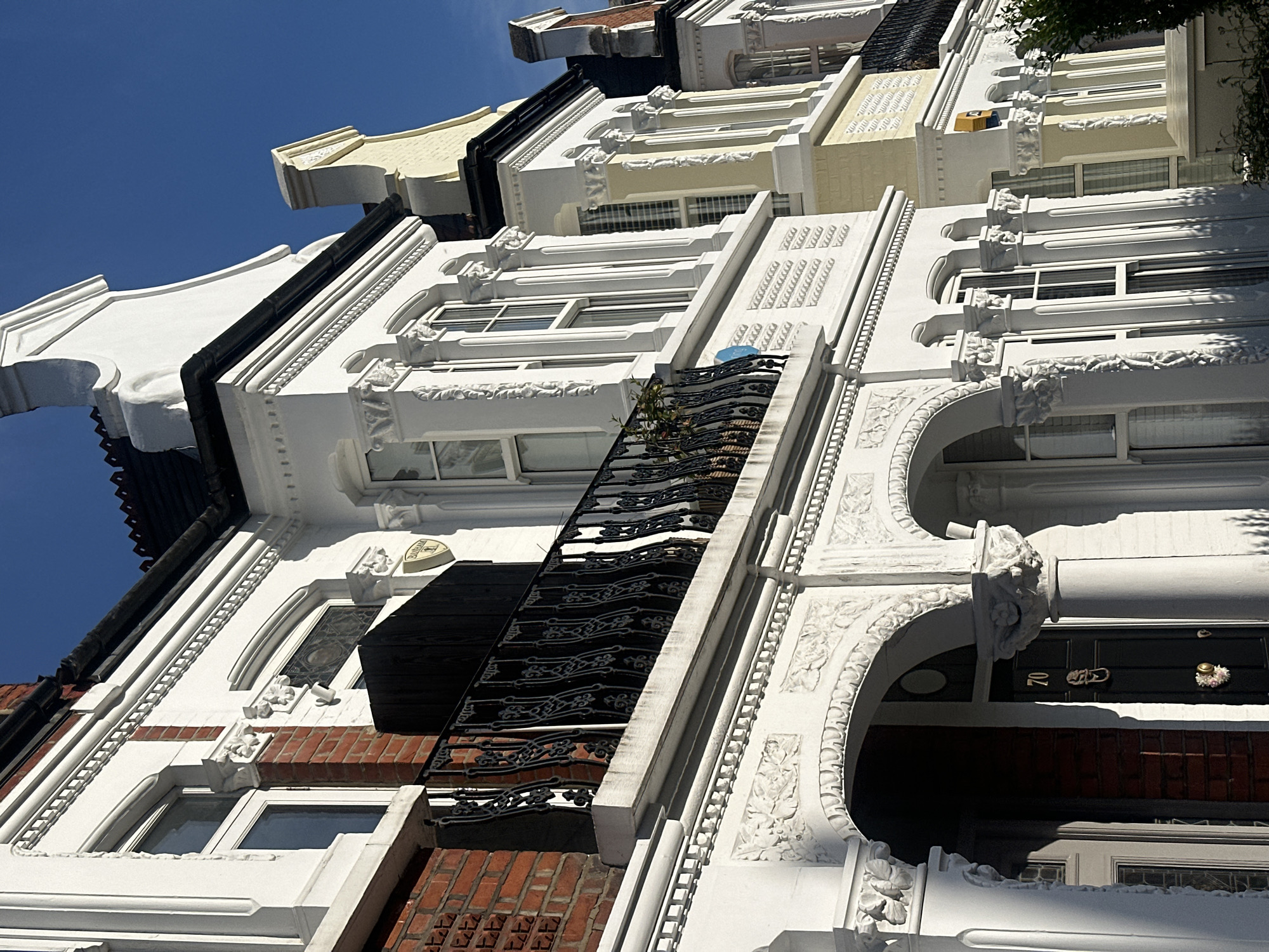The image shows a row of terraced houses in London, United Kingdom. The buildings are predominantly white with ornate facades, and some have brickwork visible. There are windows of varying sizes and shapes, with black iron balconies. The sky is clear and blue. The building on the right shows the number 70 on the door.