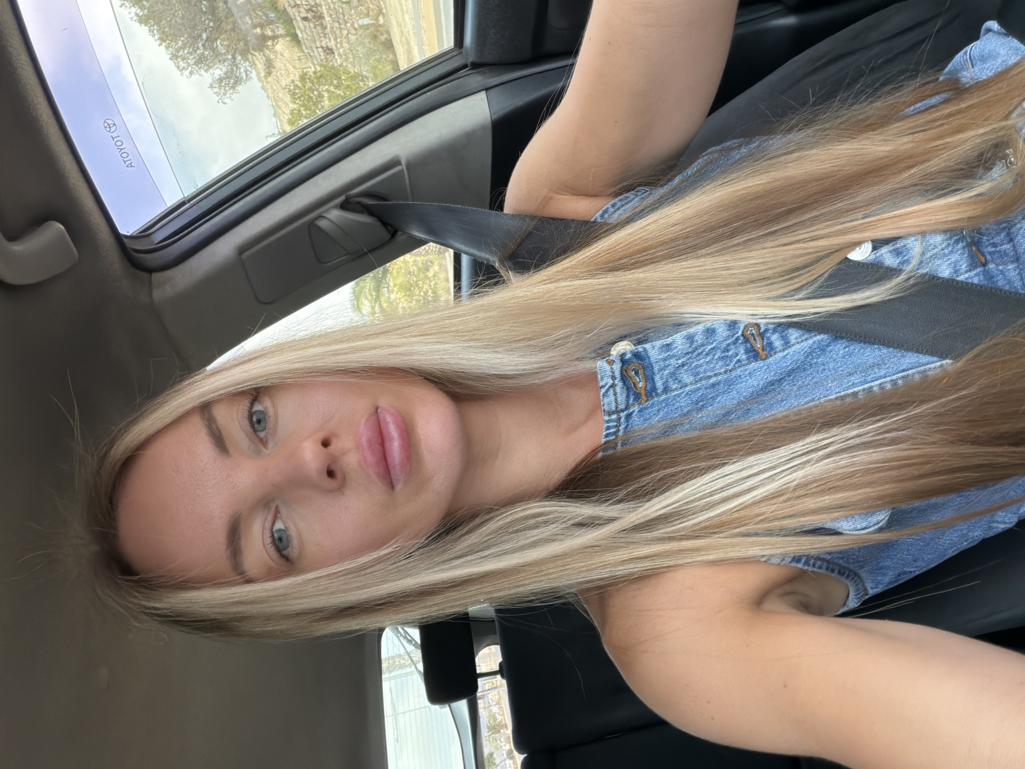 A close-up, sideways selfie shows a woman with long blonde hair and blue eyes. She is wearing a denim top and a seatbelt. The background shows the interior of a car, including the sunroof and a portion of the driver's side window, with trees and sky visible outside. The image appears to be taken during the day, with natural light illuminating the scene. The woman's expression is neutral. The word "TOYOTA" is visible on the sunroof.