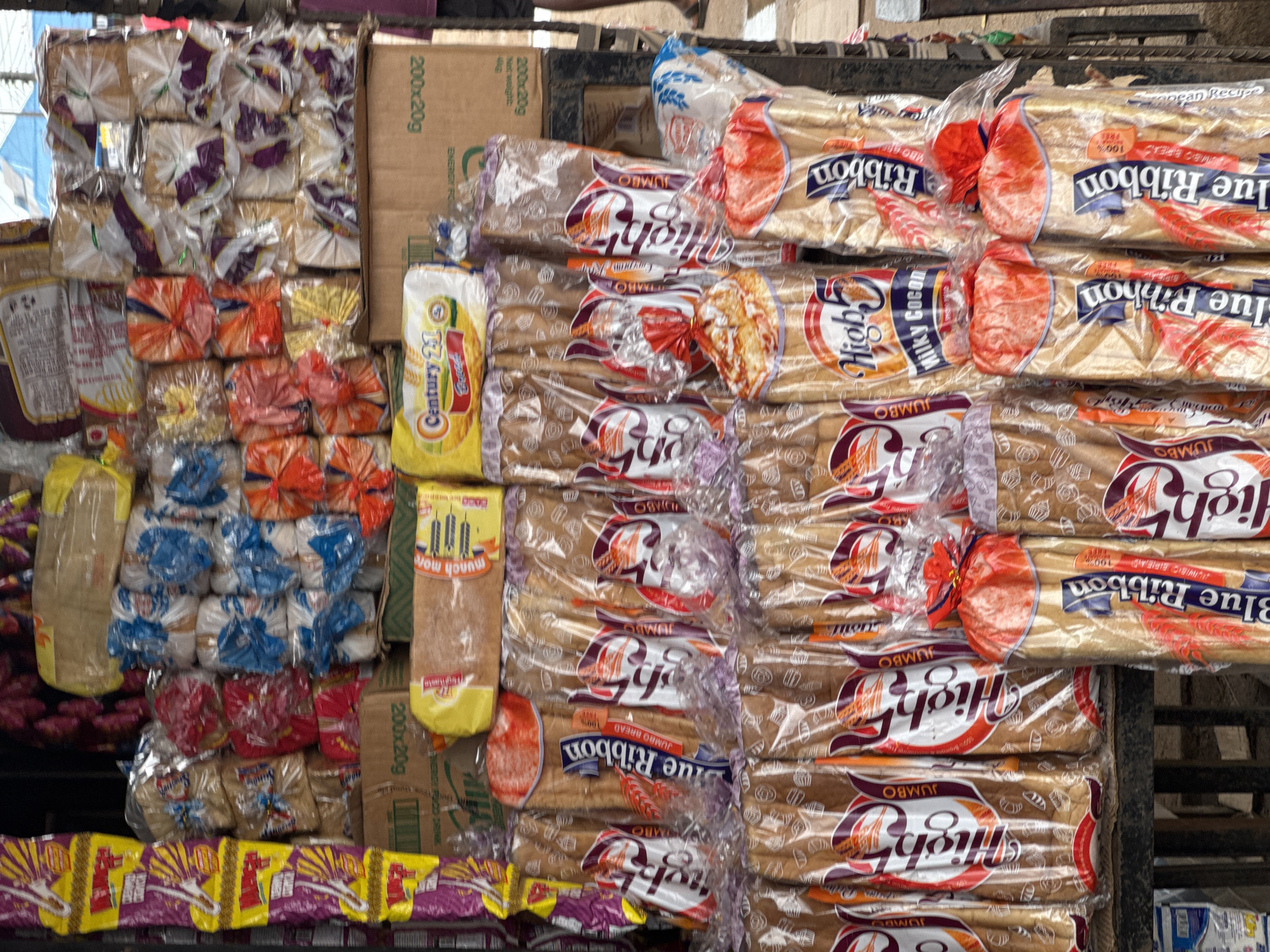 This image depicts a street-side stall in Ihiagwa, Nigeria, overflowing with various packaged food items, primarily bread. The stall appears to be constructed from simple materials, with shelves or displays holding the products. Prominently featured are loaves of bread wrapped in clear plastic, showcasing different brands and types. Some brands include "Blue Ribbon," "High 5," and "Century 21." The bread packaging displays vibrant colors and graphics, with some loaves tied with red ribbons. In the background, other packaged goods, possibly snacks or biscuits, are visible, along with some indistinct items that may be produce or household goods. The overall impression is that of a bustling local market stall offering a wide assortment of staple foods.
