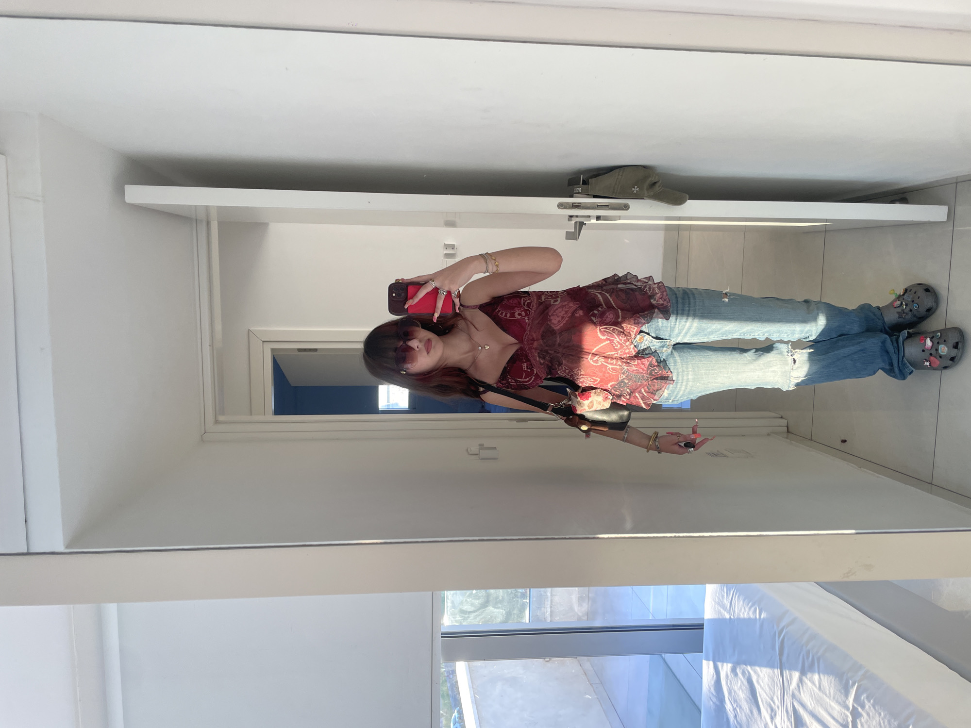 The image depicts a young woman taking a mirror selfie in a modern apartment. She is wearing a burgundy patterned camisole, distressed light-wash jeans, and grey Crocs adorned with charms. She has on large sunglasses and is holding her red-cased smartphone in her right hand. A dark handbag with a patterned scarf tied to it hangs from her left shoulder. She is also wearing several rings and bracelets. The background shows a clean, minimalist interior with white walls and tiled flooring. To the left, a large window reveals a balcony with an outdoor space and a glimpse of greenery. A white-sheeted bed is partially visible in the reflection. To the right, a door is ajar, revealing a glimpse of a blue-painted room, and a green jacket hangs on a hook near the doorframe. The overall impression is one of casual style in a bright, contemporary living space. The scene is consistent with a residential setting within the city of Paphos Municipality, Cyprus, suggested by the architectural style and bright, sunny atmosphere, often characteristic of Mediterranean locales.