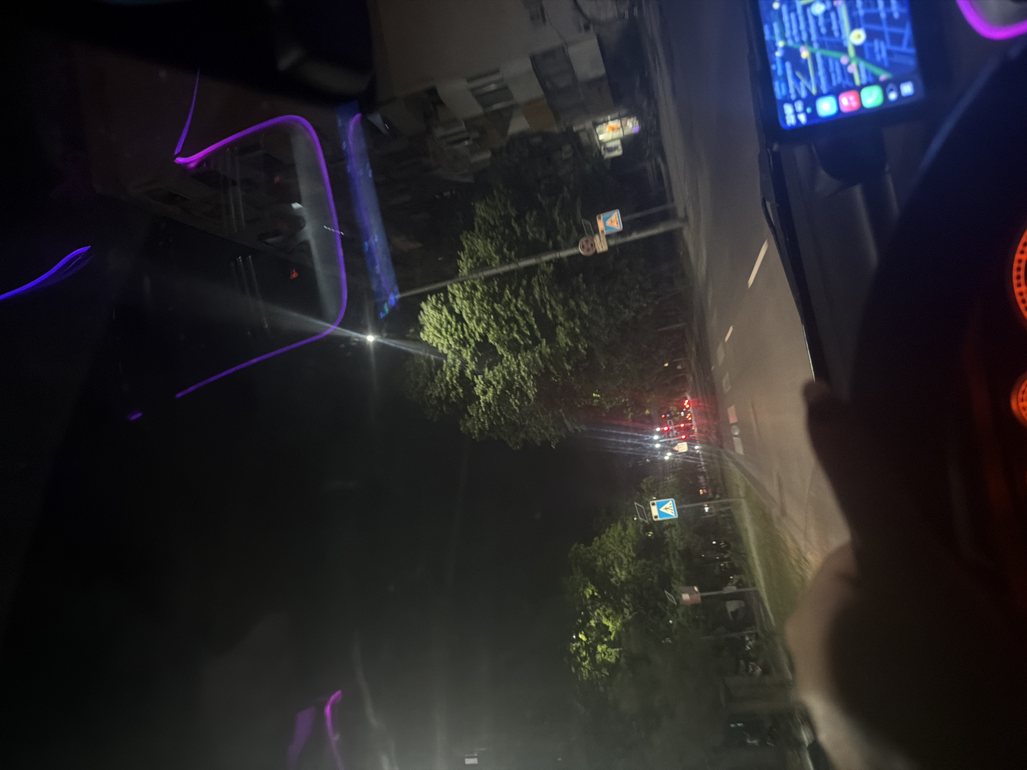 The image shows a nighttime view from inside a car, presumably the driver's perspective. The road ahead is illuminated by streetlights and traffic lights. A navigation app is visible on a screen mounted near the steering wheel. Street signs are present, indicating a typical urban environment. The purple ambient lighting inside the car creates a modern, personal feel. Based on the street signs and general urban characteristics, the location could be in Plovdiv, Bulgaria, or any similar city.