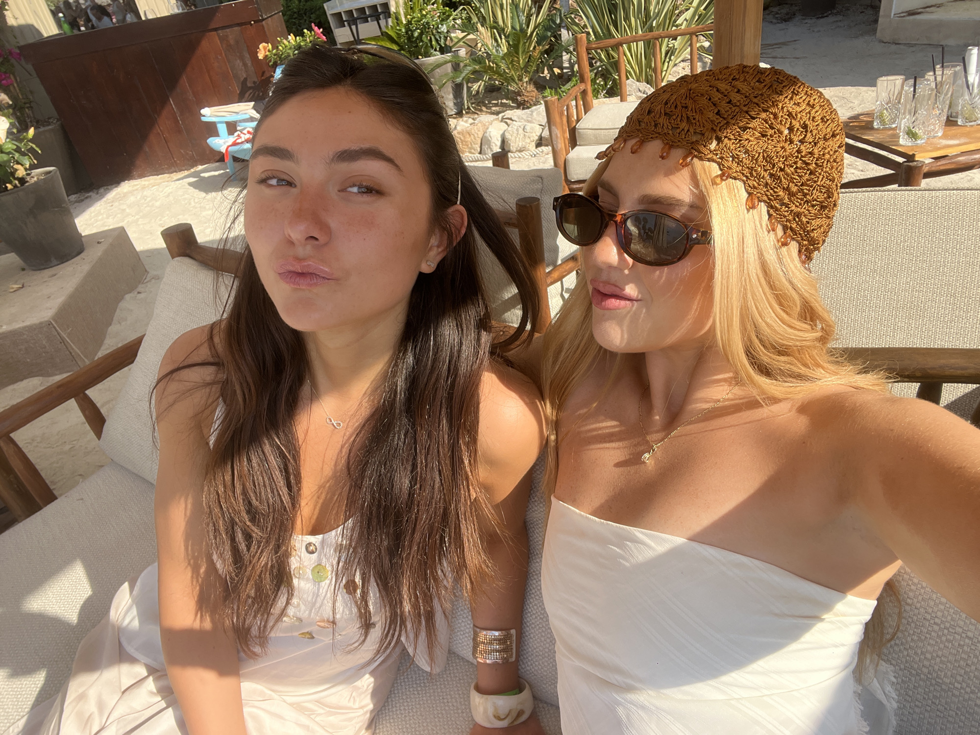 Two women are seated side-by-side outdoors on a light-colored, cushioned bench with a wooden frame. The woman on the left has long, dark brown hair and is wearing a white dress with buttons. She's making a pursed-lips expression. The woman on the right has blonde hair, sunglasses, and a crocheted head covering with beaded trim. She is wearing a white strapless top. They are both wearing necklaces and bracelets. The lighting indicates a sunny day, and the setting includes foliage, palm trees, wooden furniture, and drinks on a nearby table. The location is likely in Cannes, France, based on the typical style of vacation venues found there.