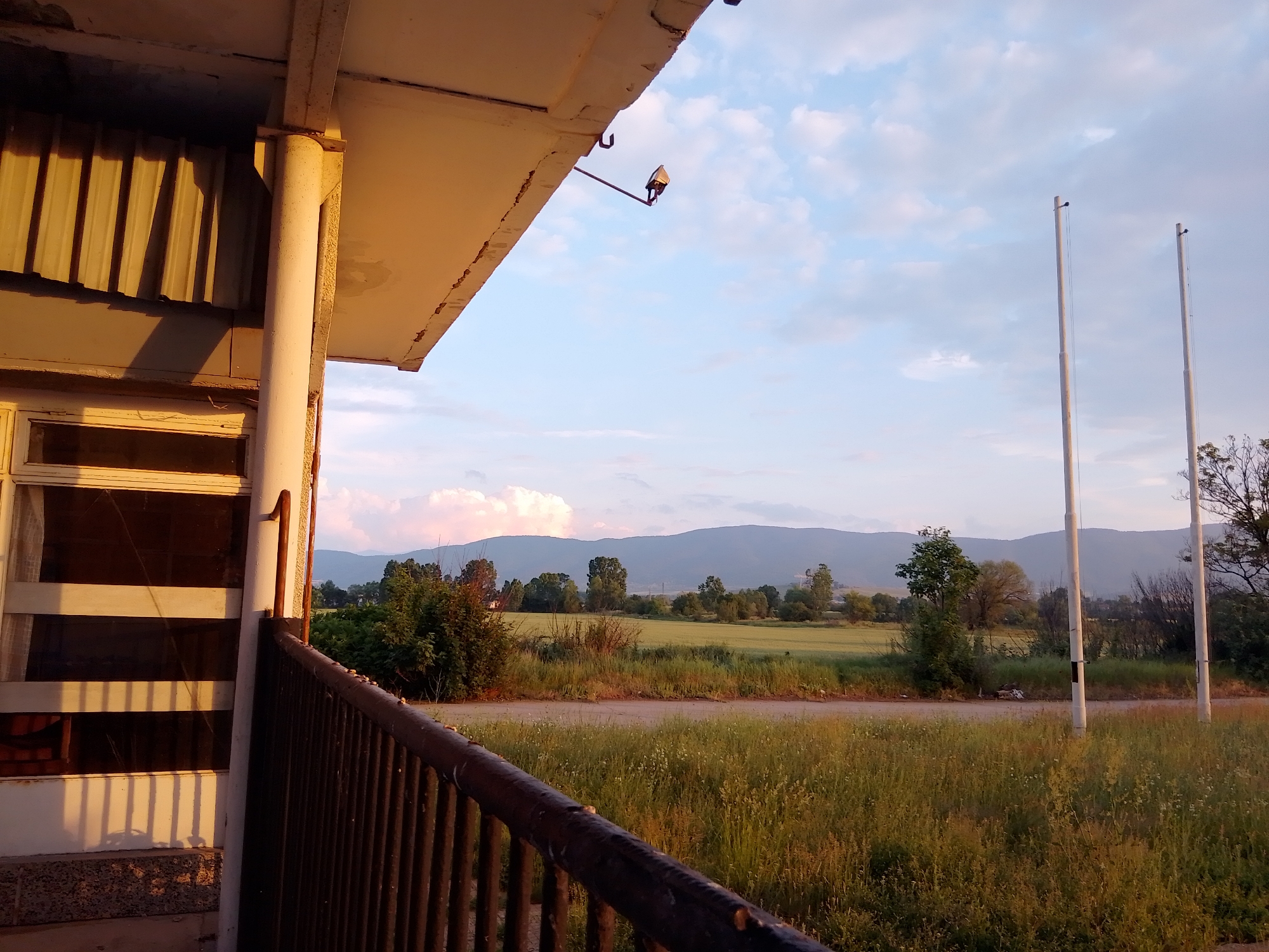 The image presents an outdoor landscape viewed from an elevated perspective, likely a balcony or porch, in Unknown, Unknown. The scene is illuminated by warm, low-angle sunlight, indicating either late afternoon or early morning, which casts long shadows.
In the immediate foreground, on the left, a section of a building structure is visible. This includes a vertical, light-colored column with visible texture and minor deterioration, and a dark metal railing with vertical bars that extends diagonally across the bottom. To the left of the column, a window or door frame with multiple horizontal divisions is partially shown. Above this, a corrugated metal section and the underside of a roof overhang, exhibiting cracks, are present. A dark, box-shaped fixture, possibly a security camera or light, is mounted on a metal arm extending from the underside of the overhang.
The midground features an unpaved road, likely dirt or gravel, traversing horizontally across the frame. This road is bordered by dense, unkempt grasses and various wild vegetation. Beyond the road, a green field, possibly agricultural, stretches out, dotted with scattered deciduous trees and bushes. On the right side of the midground, two tall, slender, light-colored poles, potentially flagpoles or utility poles, stand vertically, each topped with a dark, small cap or finial.
The background consists of a long range of hazy, bluish-grey mountains or hills. The sky above is a pale blue with scattered white, cumulus clouds; some clouds on the left display faint pink and orange hues. There are no individuals or discernible actions depicted in the image.