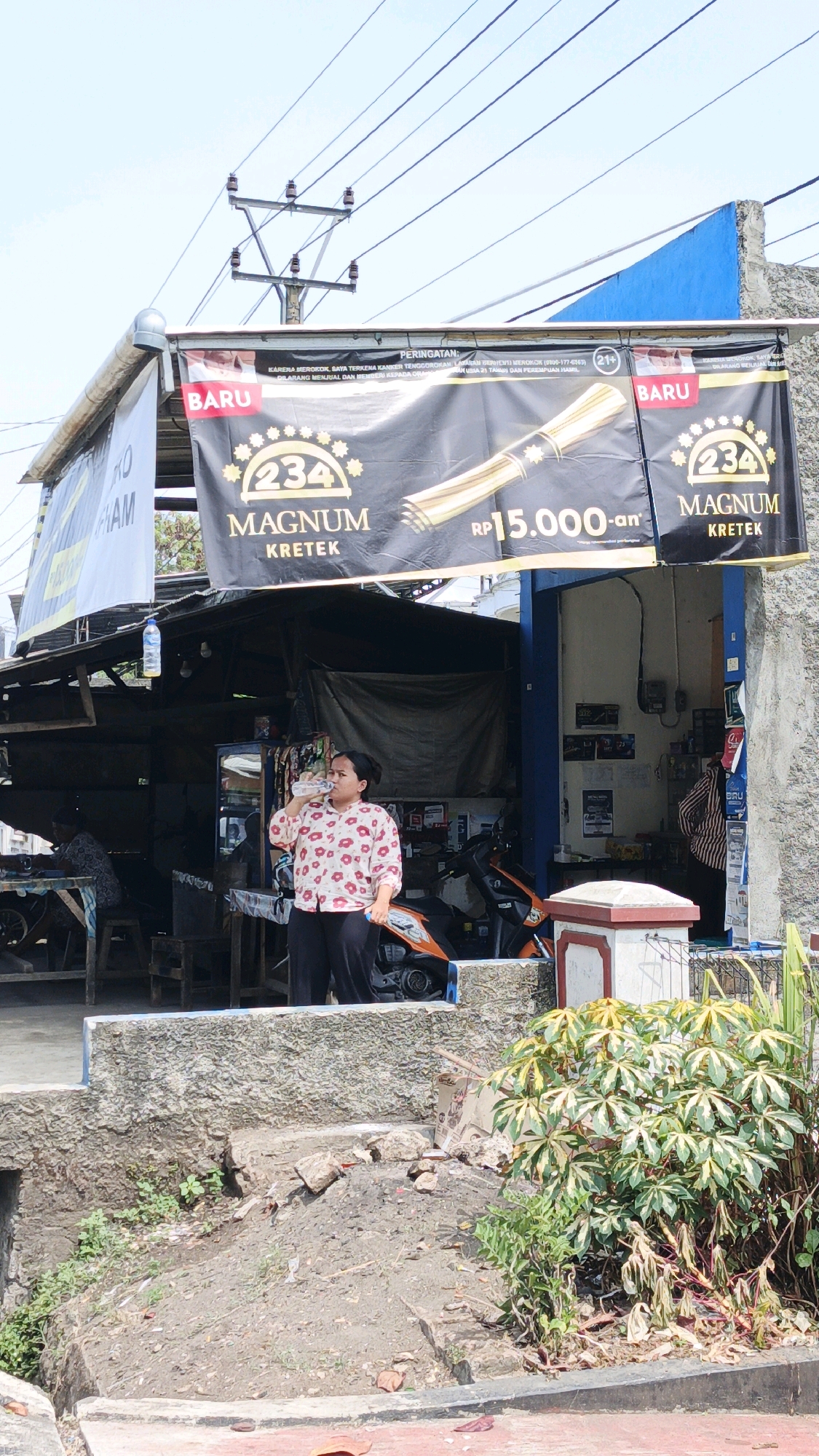In Cibenda, Indonesia, a woman in a floral shirt and black pants stands outside a small storefront, drinking from a bottle of water. Above the storefront, a black banner advertises "234 Magnum Kretek" cigarettes, with text indicating they are for individuals aged 21 and over and warning of health risks. The banner displays the price as Rp 15,000. The storefront itself appears to be a simple structure with a corrugated metal roof and an open front. Inside, various items are visible, including shelves stocked with products and a motorcycle parked to the right. The scene is set on a sunny day, with a clear blue sky and electrical wires overhead. The ground in front of the storefront is uneven, with dirt, rocks, and some vegetation.