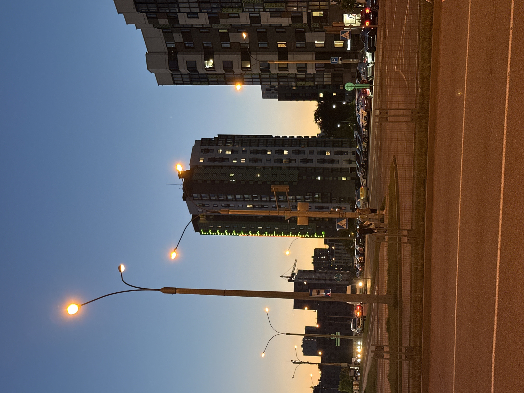This wide-angle urban scene captures a residential area at dusk or early night. The sky, transitioning from deep blue overhead to a warm orange-yellow near the horizon, dominates the upper left. Multiple modern, multi-story residential buildings line the left and center of the frame, their windows illuminated, suggesting evening activity. One prominent building in the middle distance features a distinctive long, horizontal green and yellow light display.
Numerous streetlights with warm, glowing lamps are scattered throughout, providing ample illumination. A large streetlight pole with multiple lamps stands prominently in the foreground on the left, while others recede into the distance.
On the right, a wide asphalt road or parking area is visible, marked with white lines. It is separated from a narrow grassy strip and parked cars by a low metal fence. Several vehicles, predominantly cars, are parked along the side, some displaying illuminated taillights.
Visible text and signs include a blue square 'P' sign (indicating parking), a triangular sign depicting a bicyclist, and a round green sign with a white arrow. A construction crane is faintly visible in the far background among other buildings. The scene conveys a peaceful, well-lit urban evening atmosphere in Бараўлянскі сельскі Савет, Belarus, with no specific events or clear human figures prominently displayed.