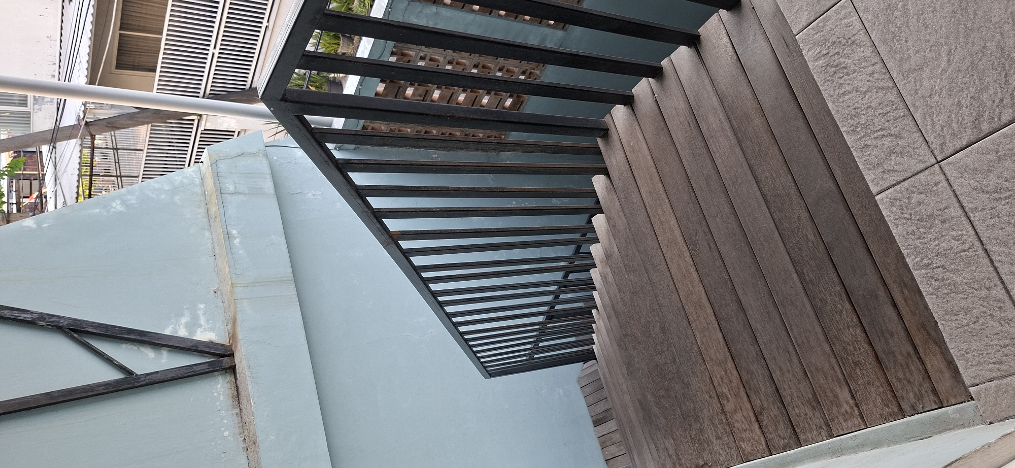 The image shows a perspective view of a wooden staircase with a dark metal railing. The staircase is positioned on an exterior of a building. The wall next to the staircase is painted a light blue-green color. In the background, there is a white metal fence and some potted plants visible. The scene appears to be outdoors. The lighting suggests it is daytime. The overall environment is clean and well-maintained. The image is taken from a slightly elevated position looking down the stairs. The city of Surabaya, Indonesia, is mentioned, but there are no specific visual cues within the image that directly confirm this location.