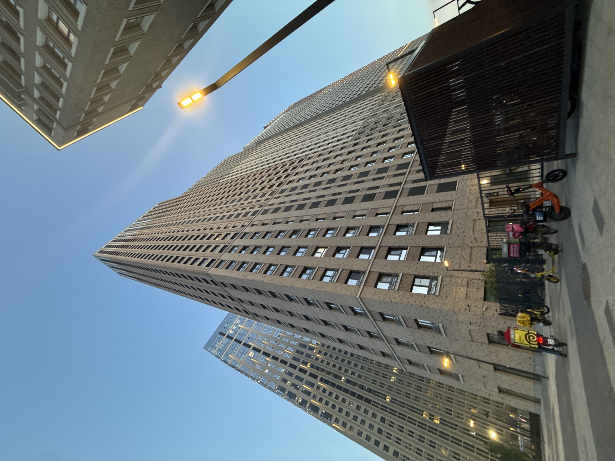 The image captures a low-angle view of a densely populated urban area in Moscow, Russia, featuring towering residential buildings with a grid-like pattern of windows. A modern street lamp with a visible light casts a warm glow towards the sky. On the right side of the frame, there's a bus stop with a slatted roof, and several electric scooters and delivery personnel on motorcycles are parked nearby, indicating an active delivery service presence. The sky is a clear blue, adding brightness to the scene.