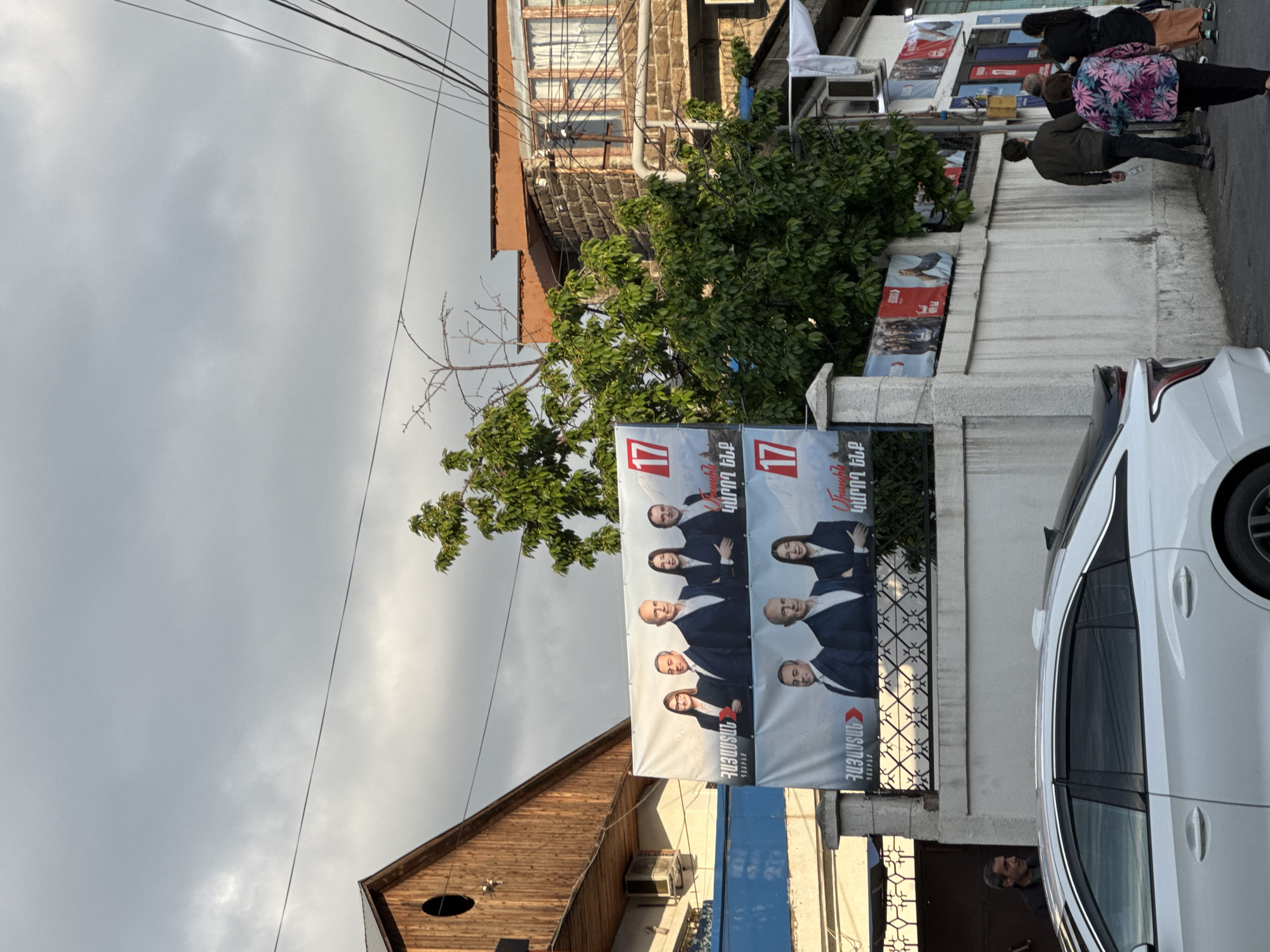 The image depicts a street scene in Yerevan, Armenia, under a cloudy sky. A political banner with the number "17" is affixed to a fence along the street. The banner features portraits of multiple people in suits and formal wear. A white car is parked next to the fence and the banner. Several individuals walk along the sidewalk in the background. Buildings of varying heights and architectural styles line the street. Overhead, electrical wires are visible. One building has a brown roof and a round window on the front.