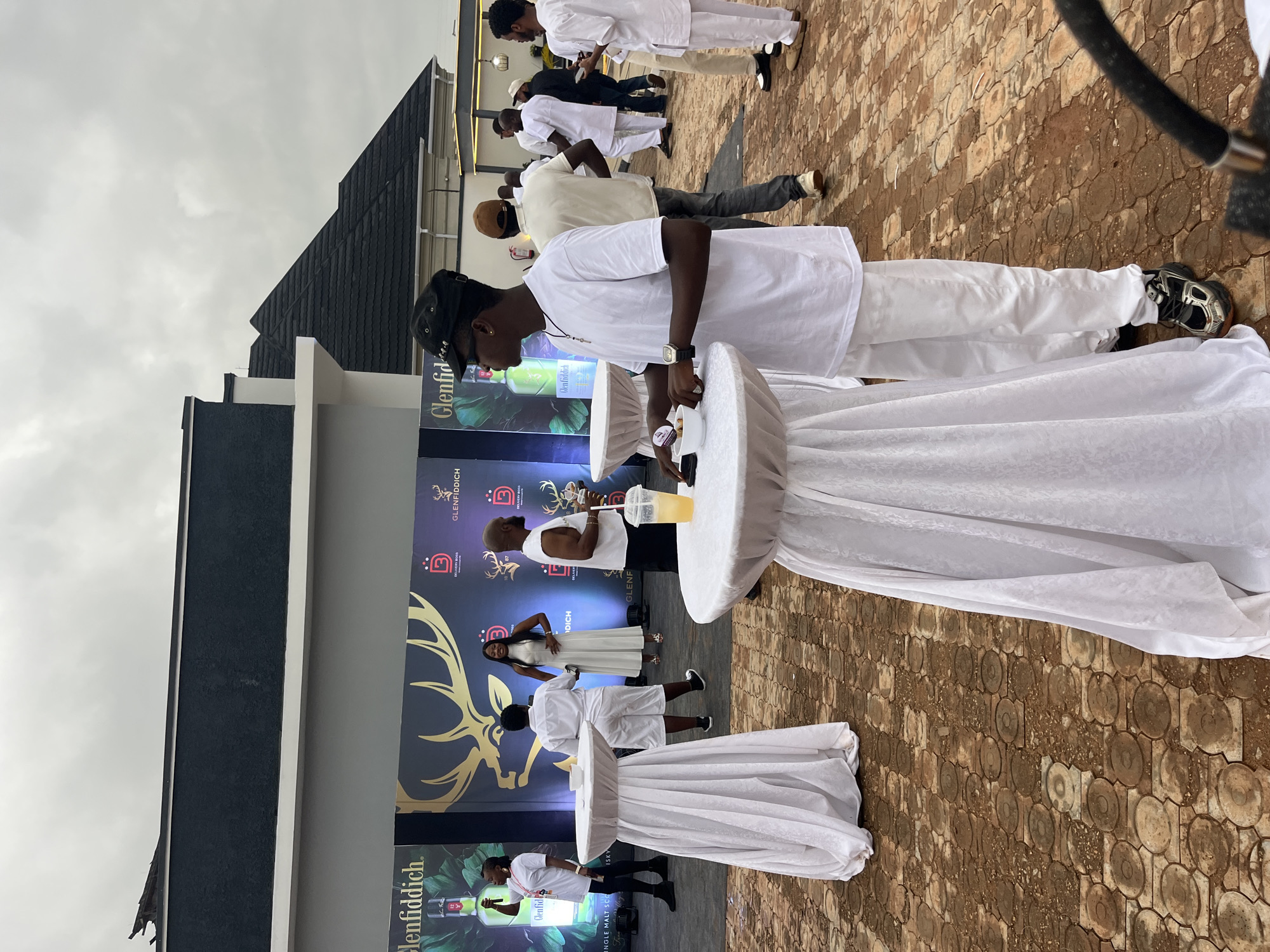 The image depicts an outdoor event, possibly a brand activation or party, with a large banner in the background featuring "Glenfiddich" branding and a stag logo. Several people are visible, dressed mostly in white attire. In the foreground, a young man wearing a white t-shirt, white pants, a black cap, and sneakers stands near a white draped cocktail table. He is looking towards the left and holding a small white bowl. Behind him, other individuals are standing and conversing. The ground is paved with patterned brown bricks. In the background, there are structures that appear to be part of a building or event setup, including a dark-colored roofline. The sky is overcast, suggesting a cloudy day. The event seems to be taking place in Bar Gada, Nigeria.