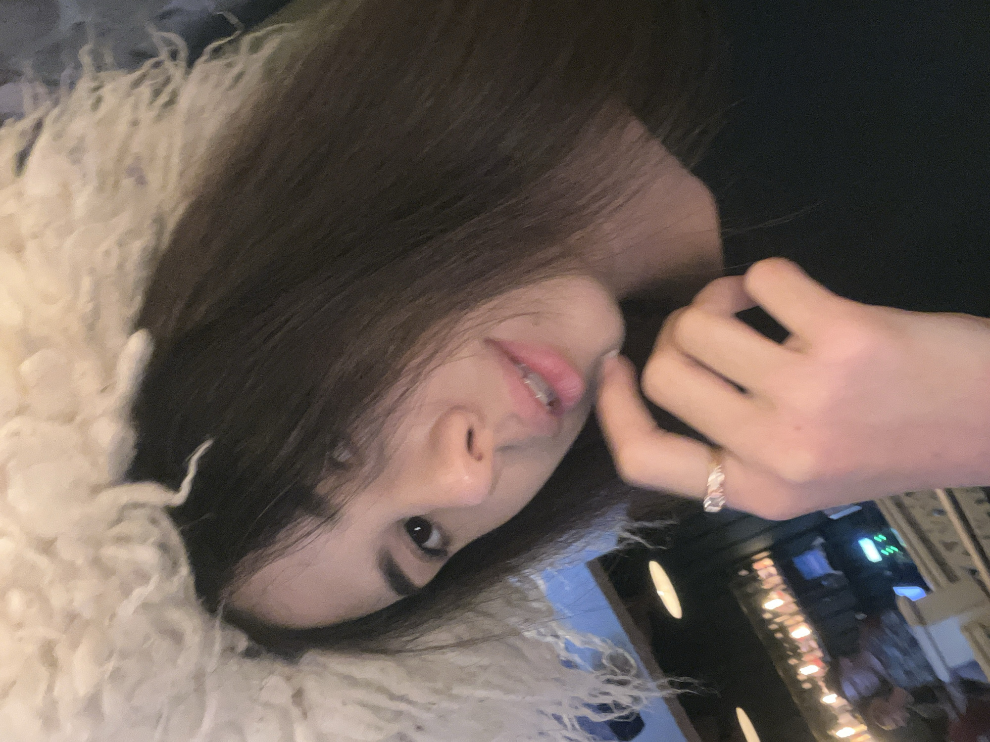 A young woman with long, dark hair is posing for a selfie. She is wearing a fluffy, white, fuzzy hat that covers the top of her head and extends down around her face. Her right hand is raised, and she is touching her chin with her index finger. She has a ring on her left hand. She is wearing a black top. The background appears to be a dimly lit establishment, possibly a bar or restaurant, with shelves of bottles visible behind her.