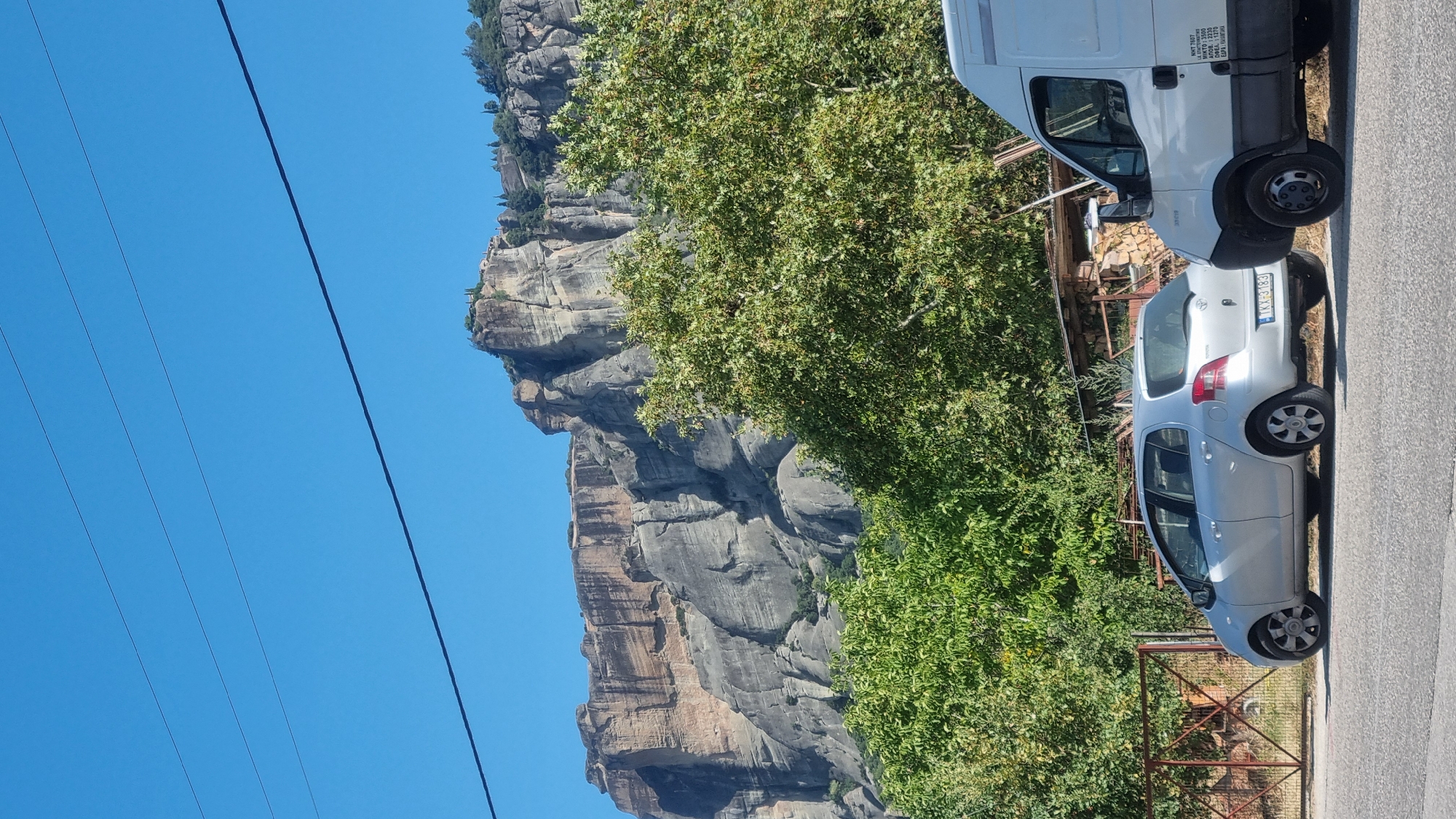 The image shows a street scene in Kalambaka, Greece, with the iconic Meteora rock formations and monasteries visible in the background. In the foreground, a silver Toyota Yaris is parked on the side of the road, its license plate reading "TKX-3183". To the right of the Yaris is a white delivery van. Behind the vehicles, lush green trees and a rustic fence are present. The sky is clear and blue. The imposing rock formations of Meteora tower over the landscape, with small structures, likely monasteries, perched precariously on their summits. The overall scene depicts a typical day in Kalambaka, a town renowned for its breathtaking natural beauty and spiritual heritage.