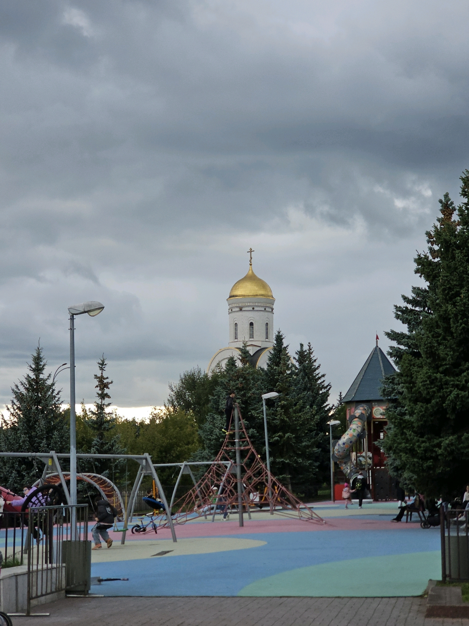 The image depicts an outdoor playground in Moscow, Russia, under a cloudy sky. The playground features various structures including swings, a climbing net, and a slide with a colorful, rocket-like design. Several children and adults are present, with some engaged in playing on the equipment and others observing. In the background, a white church with a golden dome and cross stands prominently amidst evergreen trees. Adjacent to the church, a smaller structure with a dark, conical roof is visible, also partially obscured by trees. The ground of the playground is a soft, multi-colored surface in shades of blue, pink, and green. Lampposts and metal fencing are also visible within the scene. The overall atmosphere suggests a public park or recreational area.