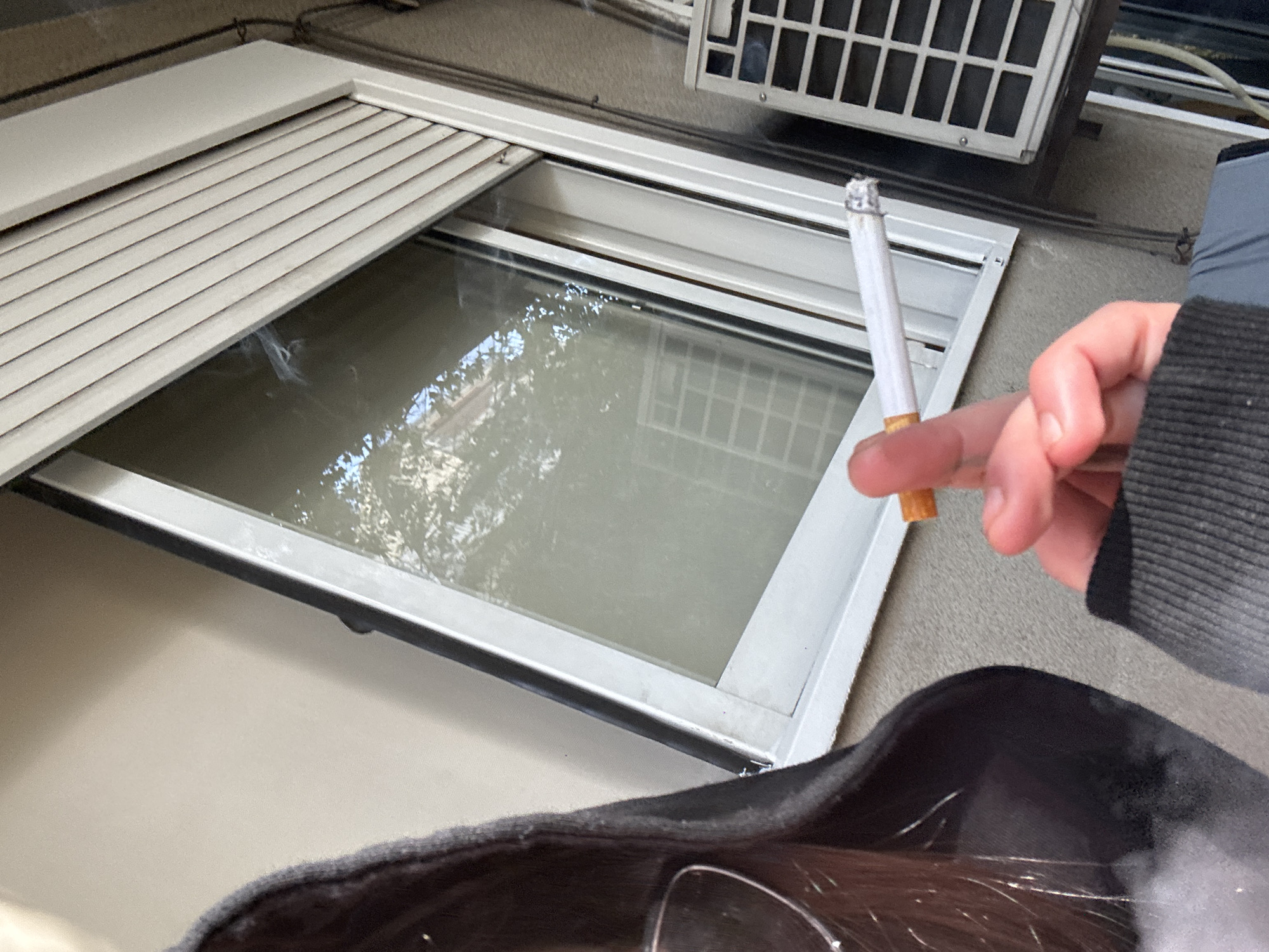 A person is holding a lit cigarette in their fingers near a window. The window has white blinds partially lowered. An air conditioning unit is mounted above the window. The setting appears to be indoors, with an outside view through the window. The location in Buenos Aires, Argentina cannot be determined from the image.