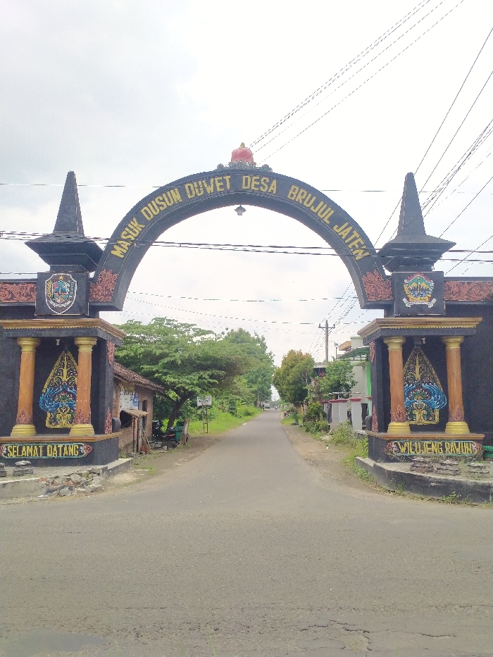An outdoor, eye-level shot captures an ornate archway marking the entrance to a village in Kecamatan Kebakkramat, Indonesia. The dark archway spans a paved road, with golden lettering on its curve that reads, "MASUK DUSUN DUWET DESA BRUJUL JATEN," translating to "Enter Dusun Duwet, Brujul Jaten Village." A decorative, reddish crown-like element adorns the peak of the arch.
The archway is supported by two robust pillars. Each pillar features a crest or emblem near its top, followed by a vibrant panel depicting traditional Indonesian wayang kulit (puppet) figures, predominantly in blue, gold, and red hues. At the base of the left pillar, the words "SELAMAT DATANG" (Indonesian for "Welcome") are displayed, while the right pillar's base reads "WILUJENG RAWUH" (Javanese for "Welcome").
The asphalt road, showing some minor cracks in the foreground, extends straight through the archway into the distance, appearing to be a single lane in each direction. It is flanked on both sides by abundant green trees and foliage, with glimpses of simple houses or structures nestled within the greenery. Overhead power lines are visible against the bright, overcast sky, suggesting it is daytime. The scene is static, devoid of people or active events, portraying a typical entrance to a rural or semi-rural Indonesian settlement.