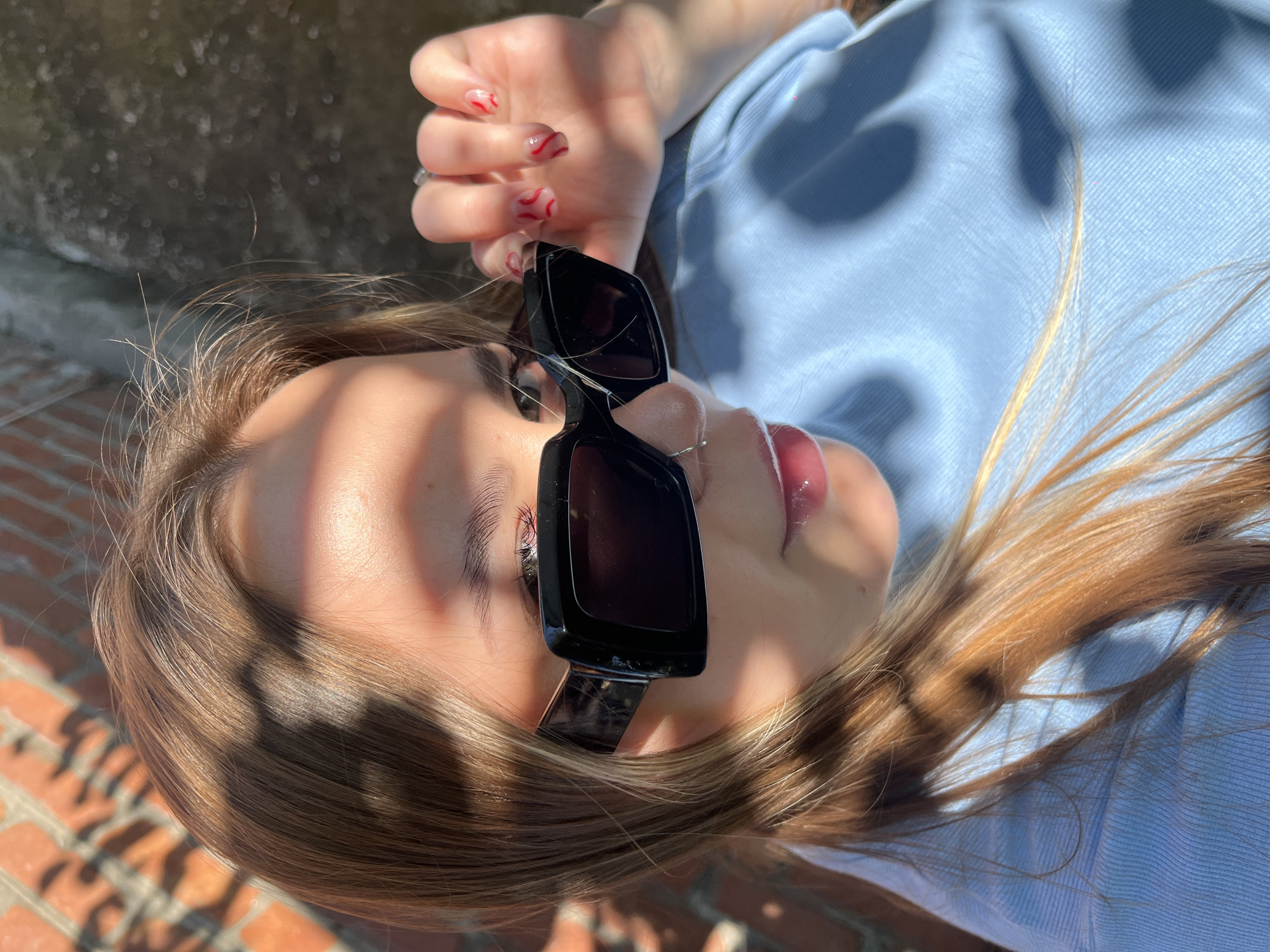A close-up, high-angle shot captures a young person reclining outdoors in Novopokrovskaya, Russia, bathed in dappled sunlight. The subject, likely female, has light brown hair cascading around their head, with some strands highlighted by the sun. They are wearing a light blue shirt or top.
The person's face is partially obscured by large, rectangular black sunglasses worn low on the nose, revealing one eye with long eyelashes above the frame. Their lips are glossy, suggesting lip gloss. One hand is visible near their face, featuring clear nails with distinctive red, wavy nail art and a ring on one finger.
Harsh, elongated shadows, possibly cast by leaves or an architectural element, create a striking pattern across the person's face, hair, and clothing, indicating bright daylight. In the background, hints of a rough, dark surface and a textured brick-like wall are visible, suggesting an outdoor setting. The overall mood is relaxed and stylish, with the strong sunlight creating dynamic light and shadow play. There is no visible text in the image.