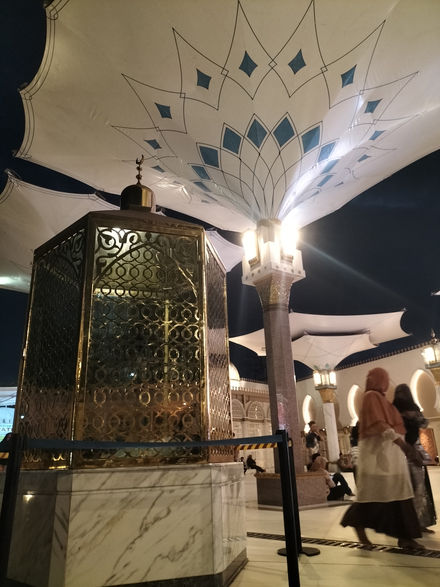In Blitar, Indonesia, at night, a golden, ornate structure, possibly a monument or a shrine, stands prominently on a marble base. The structure is enclosed by a metal grille with intricate circular patterns. Above, large white fabric canopies, decorated with blue diamond shapes and dark lines, are illuminated by overhead lights, creating an umbrella-like effect. A tall, fluted column with ornate gold accents supports one of these canopies and is lit from within. In the background, several people are visible, some walking and others seated, dressed in traditional Islamic attire. The environment appears to be an open courtyard or public space, possibly part of a religious site.